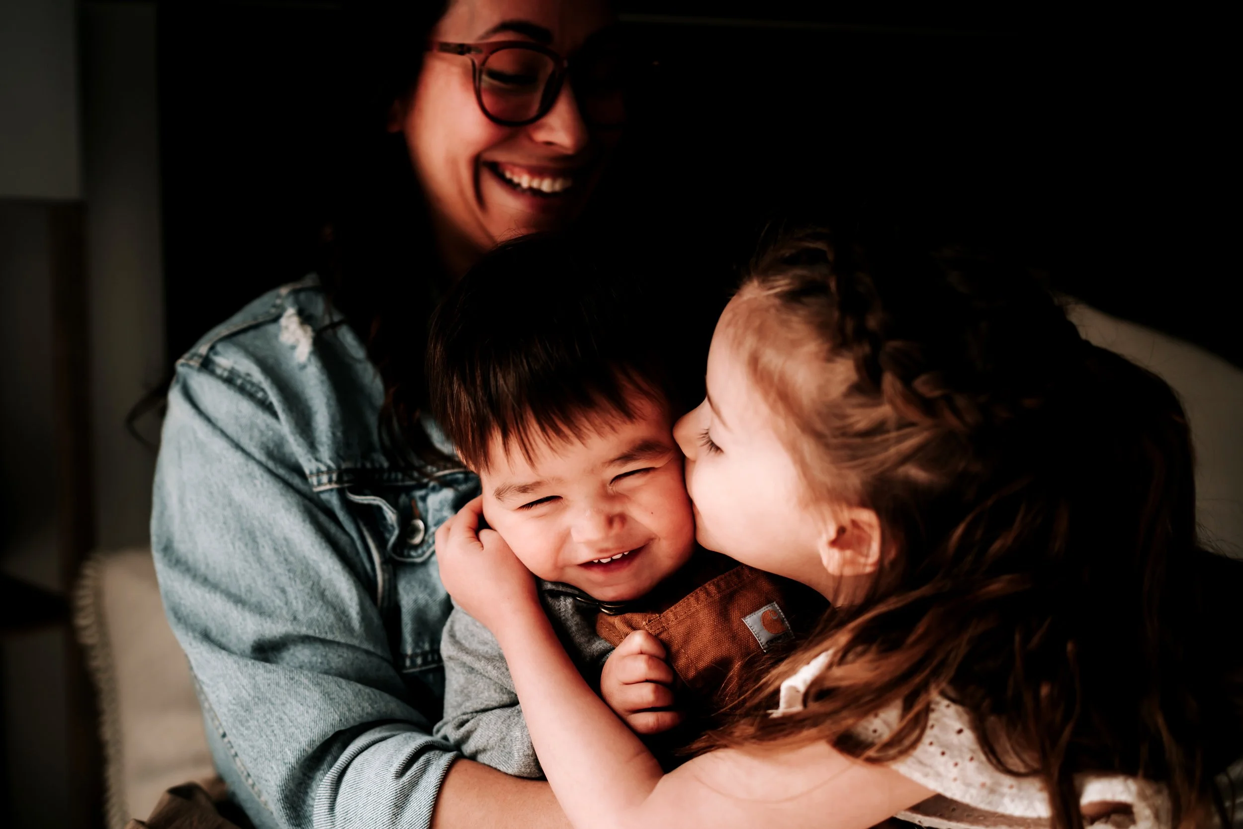 A woman with glasses and a denim jacket is holding two children, one boy and one girl, in a warm embrace. The girl is giving the boy a kiss on the cheek, and they all appear happy and smiling.