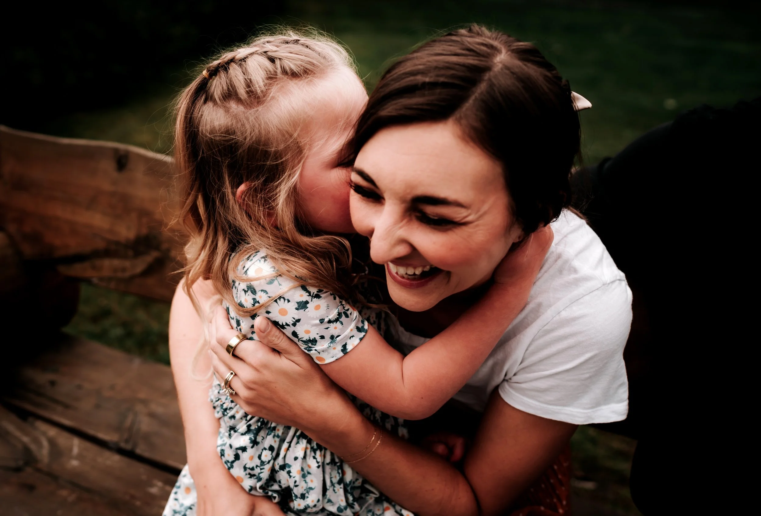 A young girl with blonde hair giving a kiss on the cheek to a woman with dark hair, who is smiling and hugging her tightly, sitting on a wooden bench outdoors.