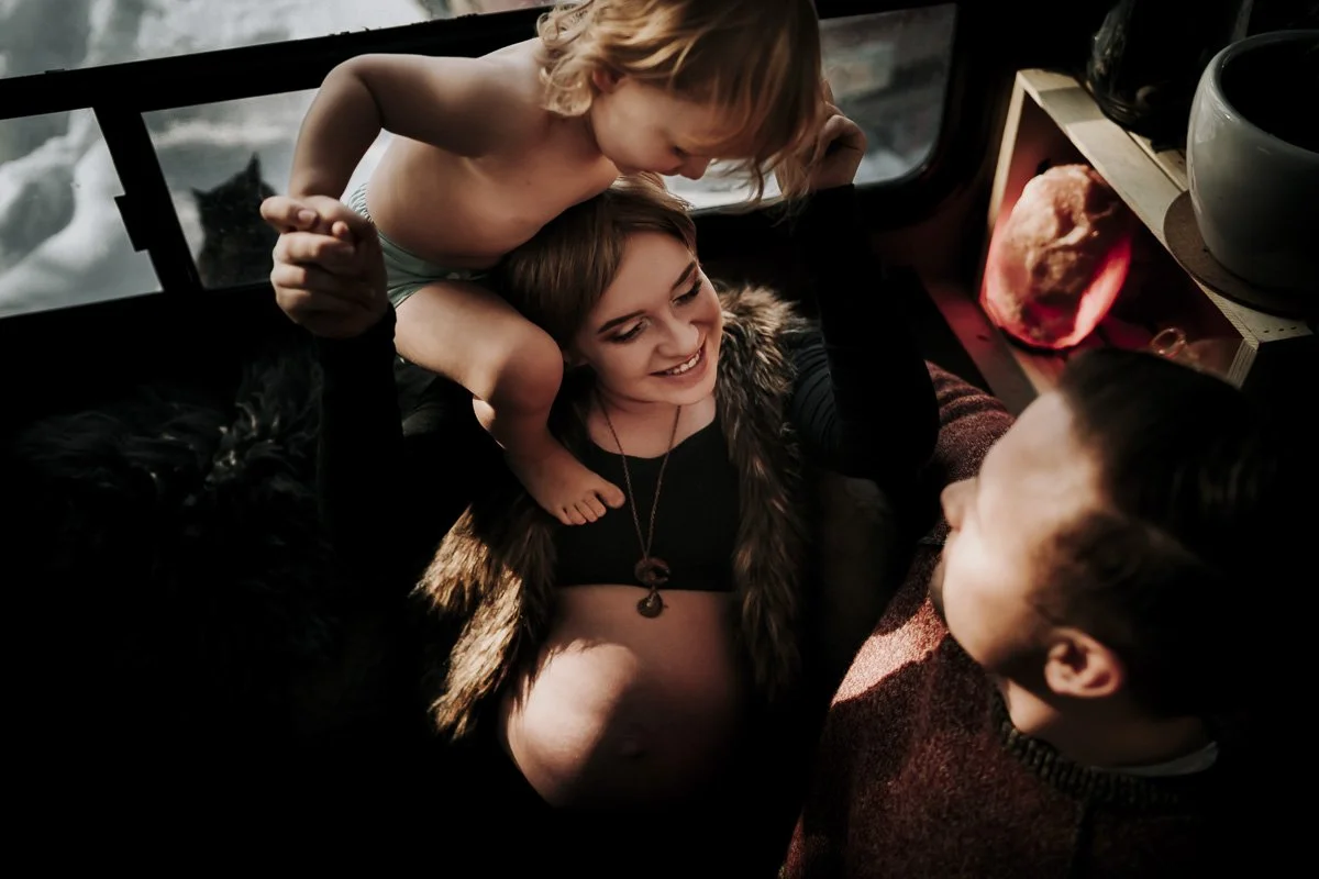 A smiling pregnant woman with two young children, one on her shoulders and one lying in front, in a cozy indoor setting with a window showing water and rocks outside.