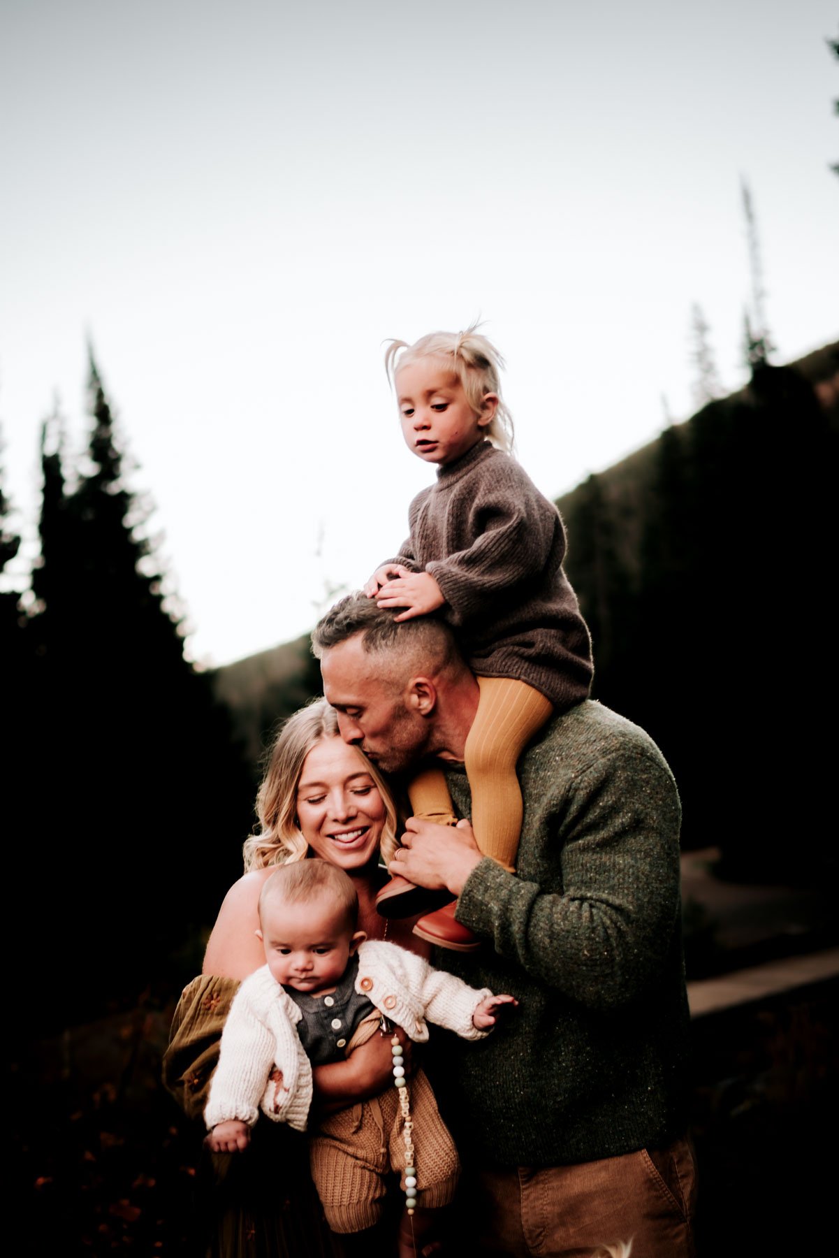 A family of five outdoors with a scenic background, with the father giving a kiss to the mother and the children around them.