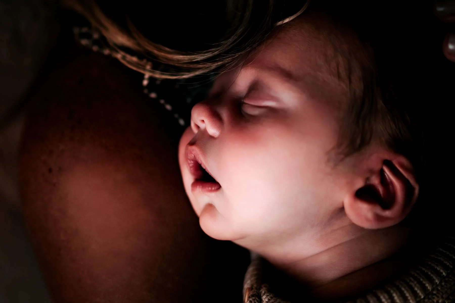 Close-up of a sleeping baby's face, with closed eyes, slightly open mouth, and light skin, resting against a person's shoulder.