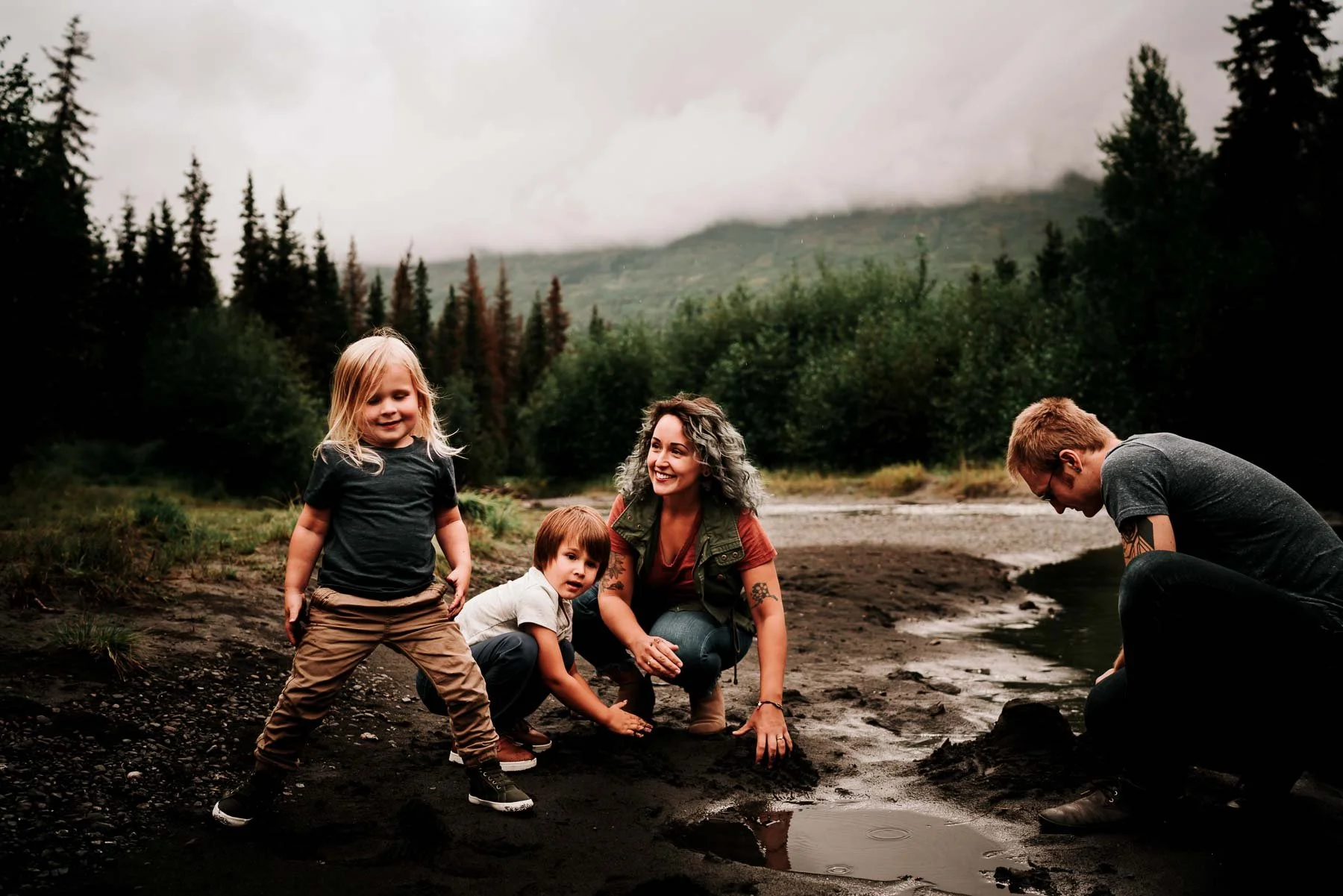 A woman and three children exploring a muddy area near a river in a forested mountainous landscape.