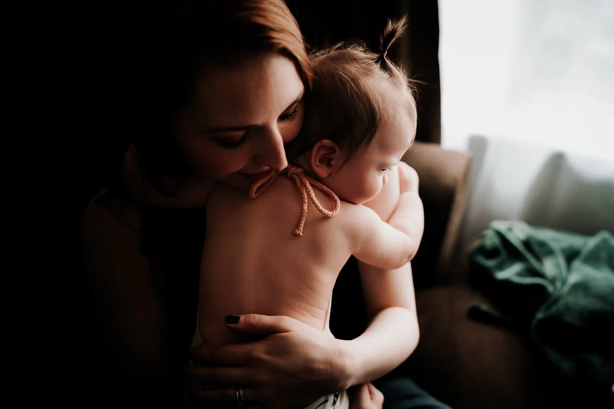 A woman holding a young child with light skin, short hair tied in a small ponytail, in a cozy indoor setting near a window, sharing an affectionate moment.