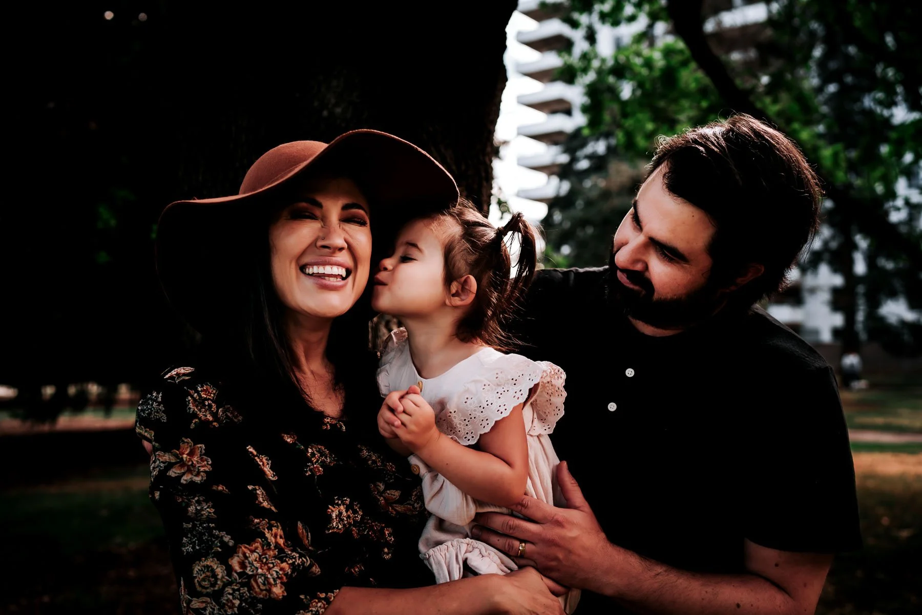 A family of three joyfully sharing a moment outdoors. The mother, wearing a large-brimmed hat and a floral top, is smiling widely. The young girl, dressed in a white embroidered dress with her hair in pigtails, is giving her mother a kiss on the cheek. The father, with dark hair and a beard, is holding the girl and looking at her lovingly. They are standing near a tree with city buildings and greenery in the background.