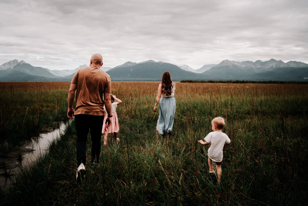 Family walking through a grassy wetland with mountains in the background under a cloudy sky.