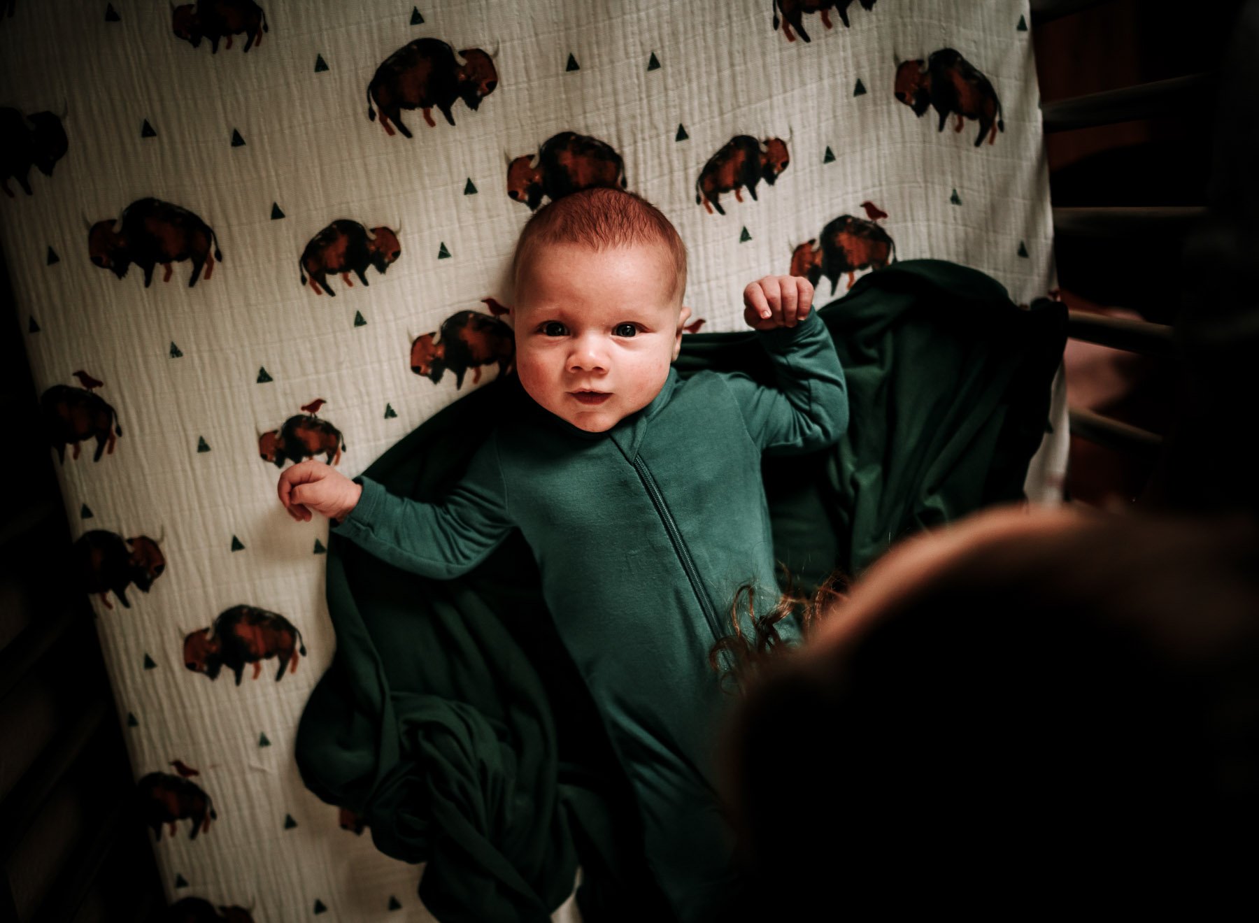 Baby lying on a bed with an animal print sheet, looking up at the camera with arms raised.