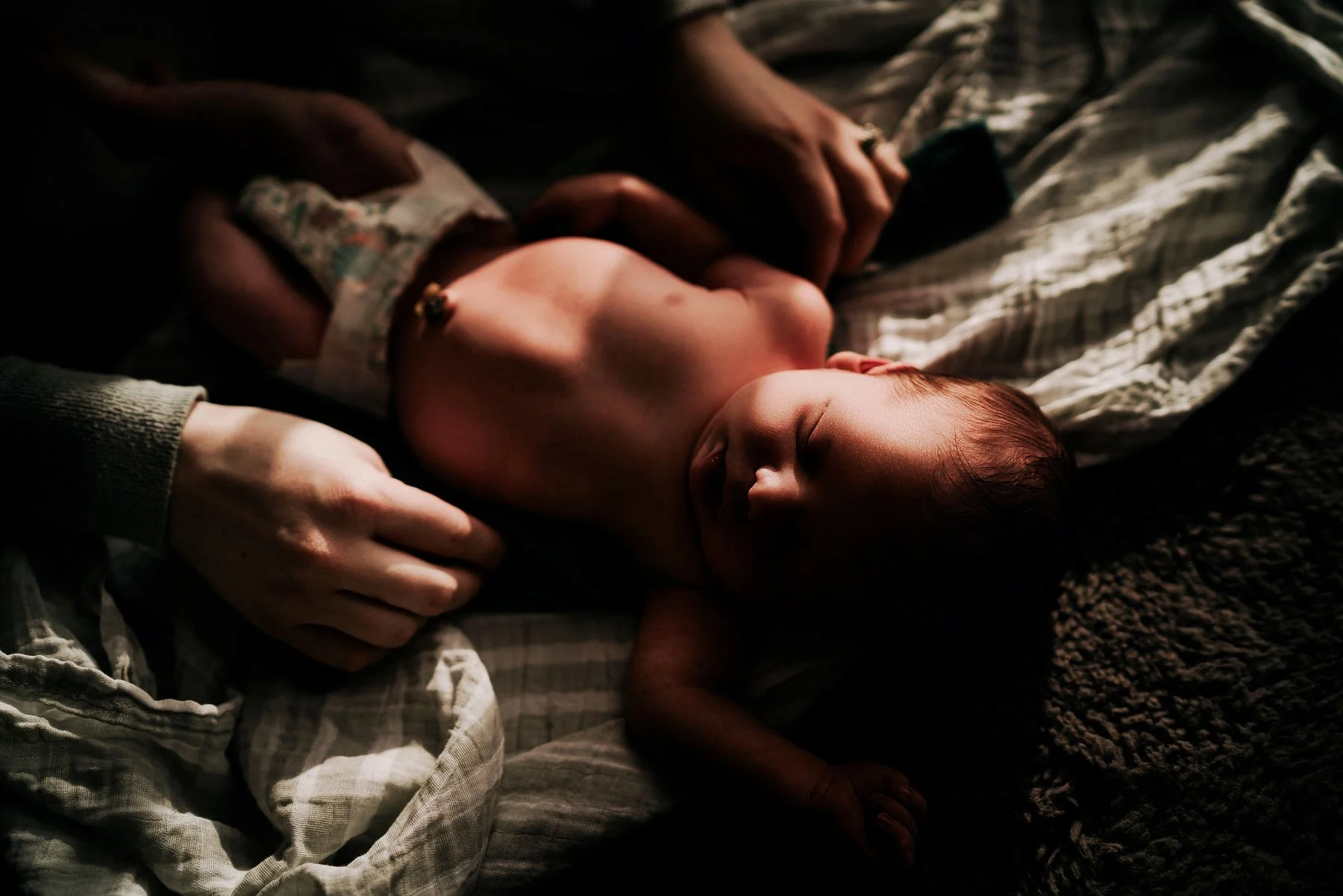 A close-up photo of a sleeping baby lying on a bed with dark blankets and bedding, with shadows cast on the baby’s face, showing a peaceful expression.