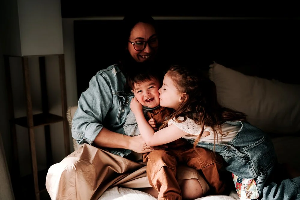 A woman sitting on a bed smiling as two children, a boy and a girl, hug and kiss each other. The girl is kissing the boy on the cheek, and they look happy.
