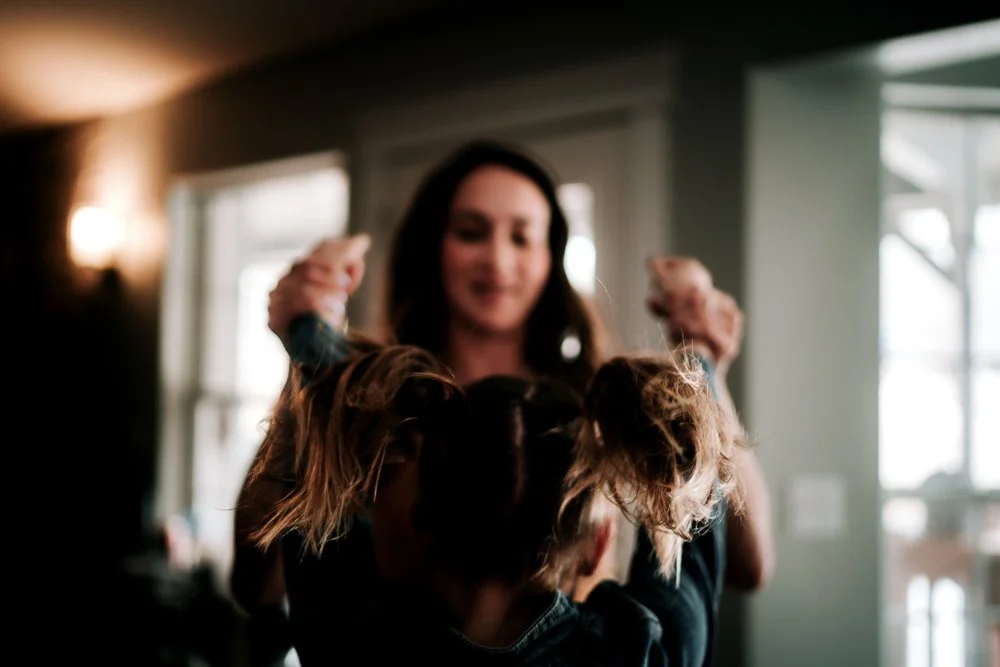 A woman smiling as she holds up two children with messy hair, in a well-lit indoor setting.
