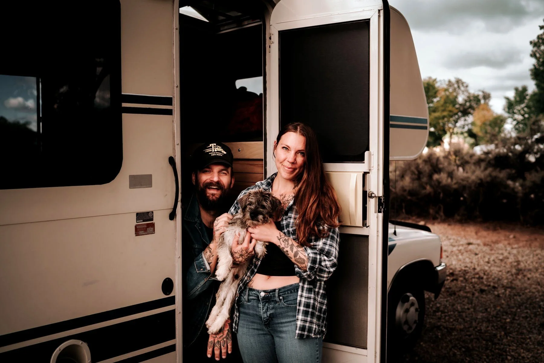 A man and woman standing in the doorway of an RV, holding a small puppy, with trees and a cloudy sky in the background.