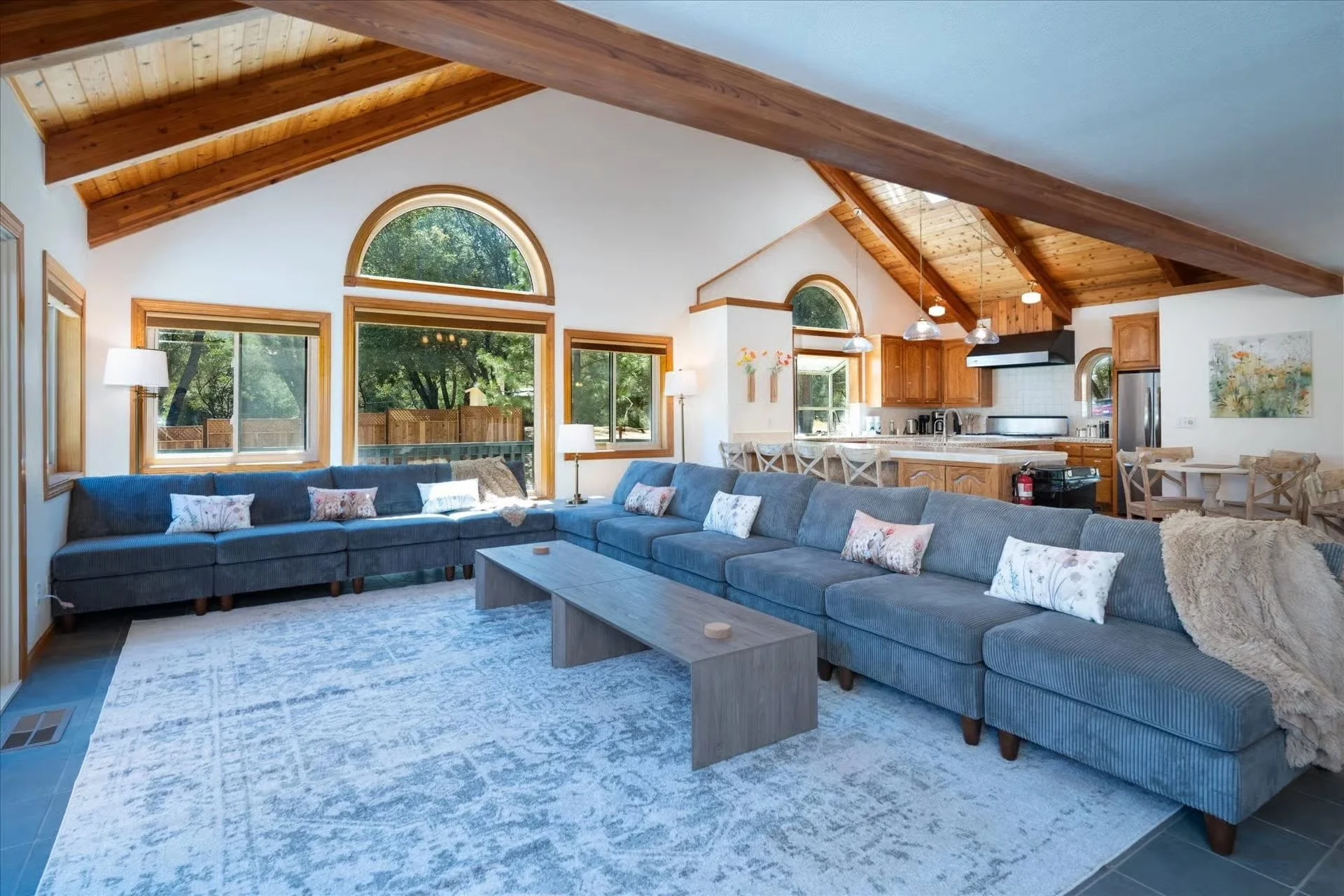 Living room with vaulted wooden ceiling, large arched and rectangular windows, blue sectional sofa with decorative pillows, white walls, wooden furniture, carpeted floor, and kitchen area in the background.