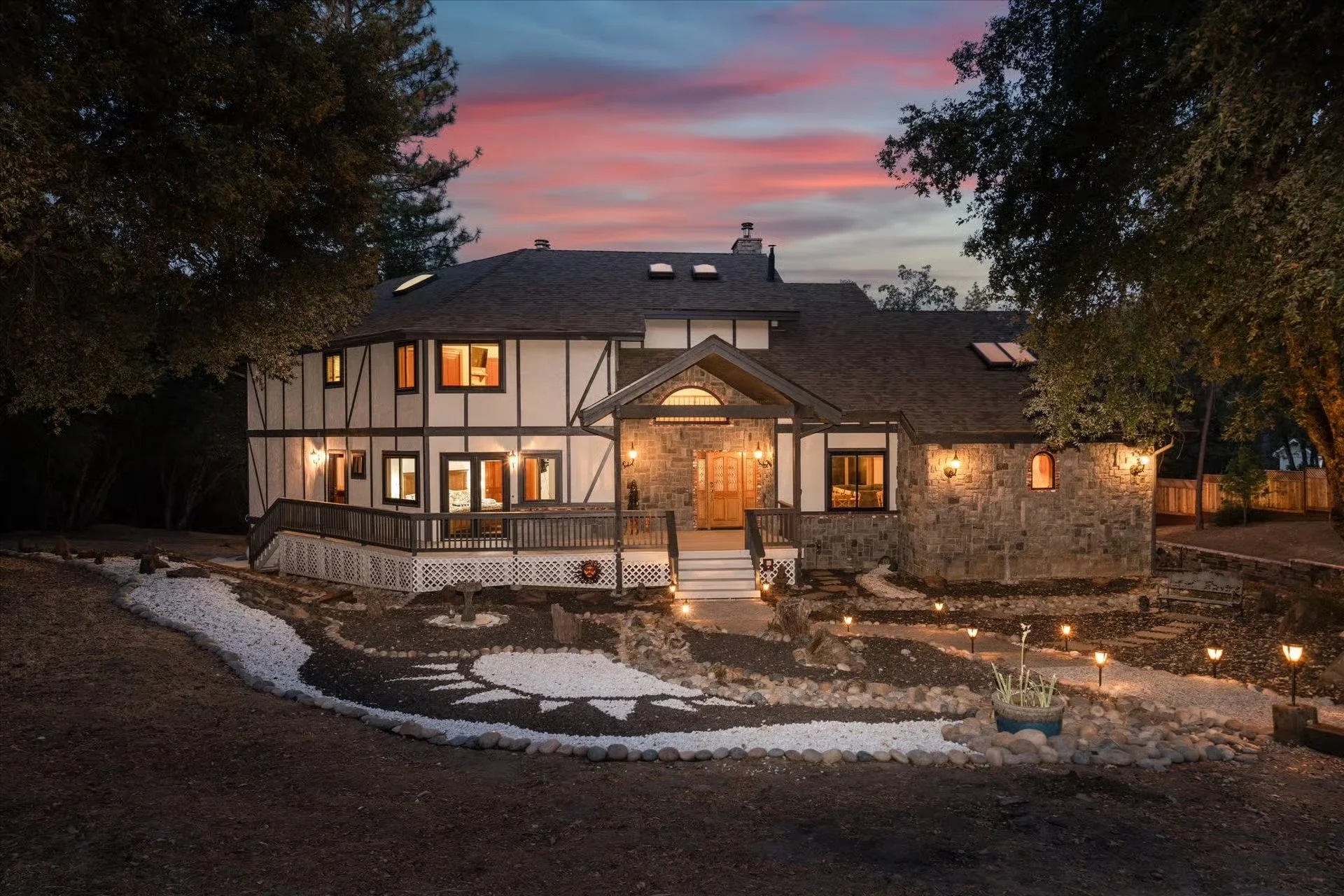 A large house with a dark roof and white and stone walls, illuminated by exterior lights. The house has multiple windows with warm light inside, a front porch with steps, and is surrounded by a landscaped yard with gravel, stones, and small path lights. Tall trees frame the house against a colorful sunset sky with pink and purple clouds.