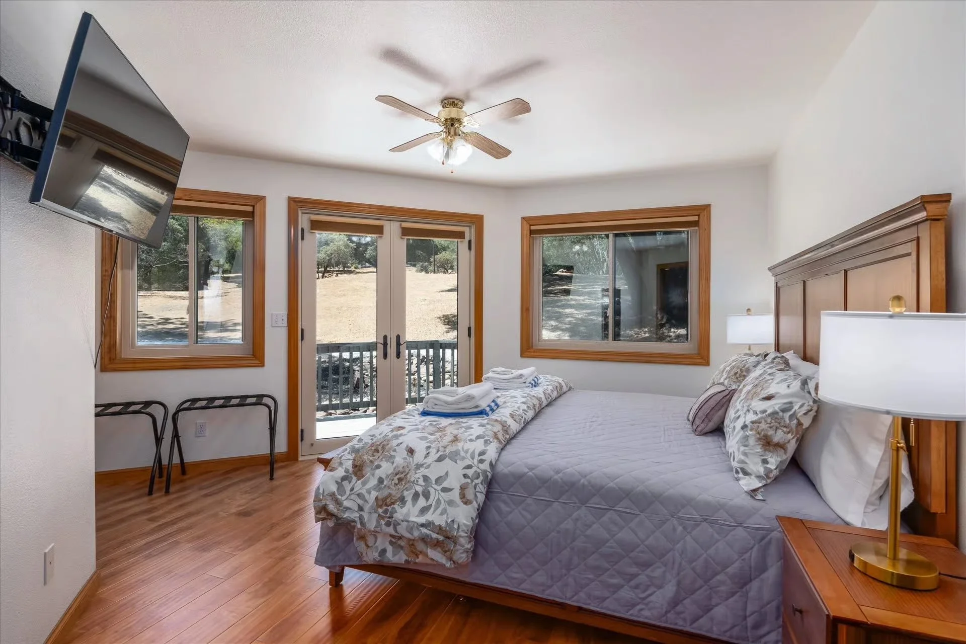 A bedroom with a bed, nightstand, lamps, windows, and a ceiling fan, overlooking an outdoor area with trees.