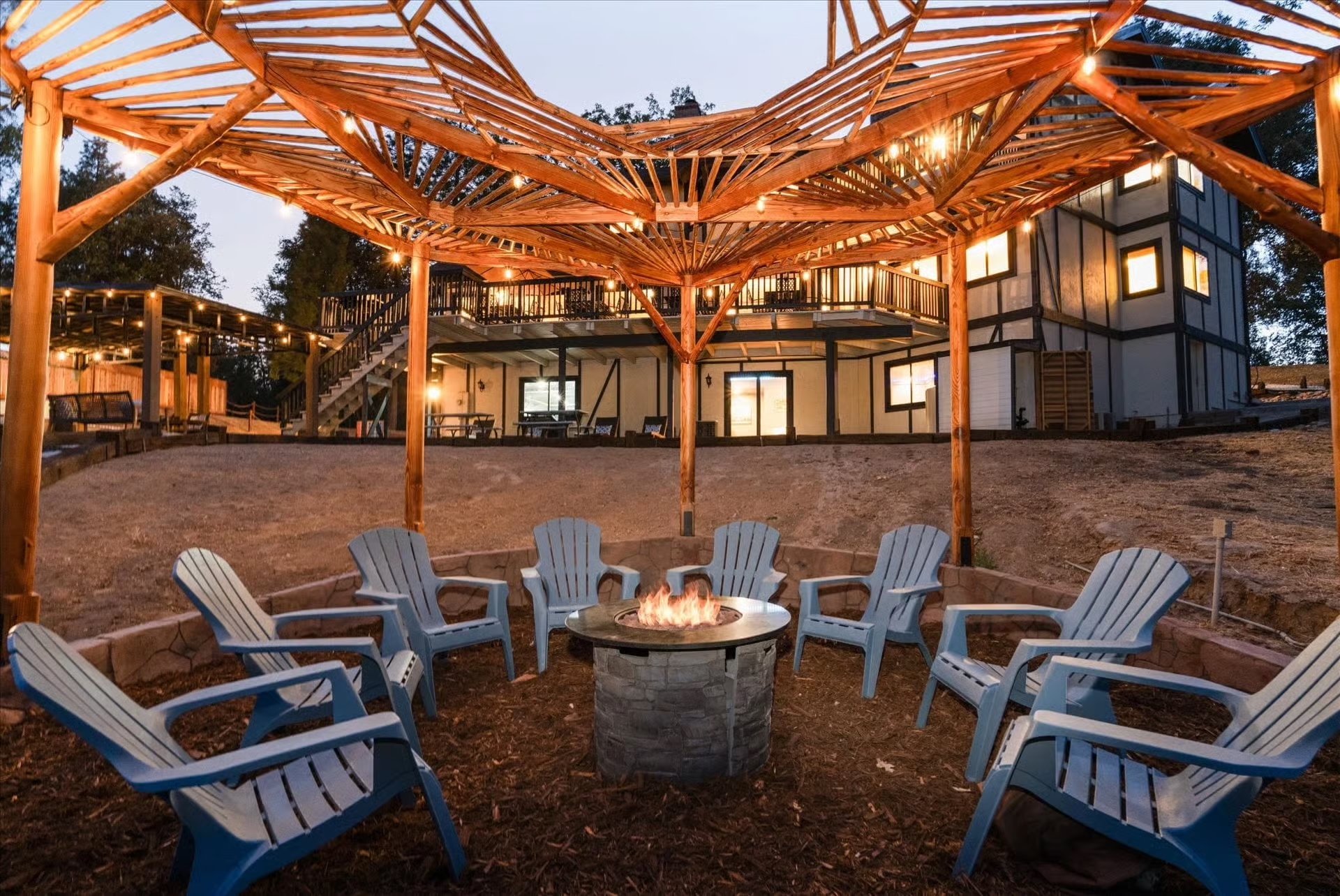 Nighttime outdoor scene with a fire pit surrounded by blue chairs, and a wooden pergola overhead. In the background, a two-story house with lit windows and an outdoor stairway is visible.
