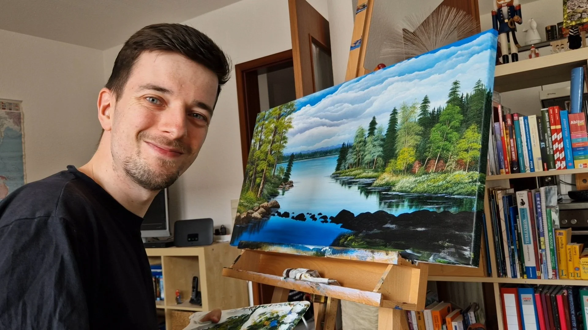 Künstler Tobias Nolte sitzt lächelnd vor einem Acrylgemälde mit einem See und Bäumen.