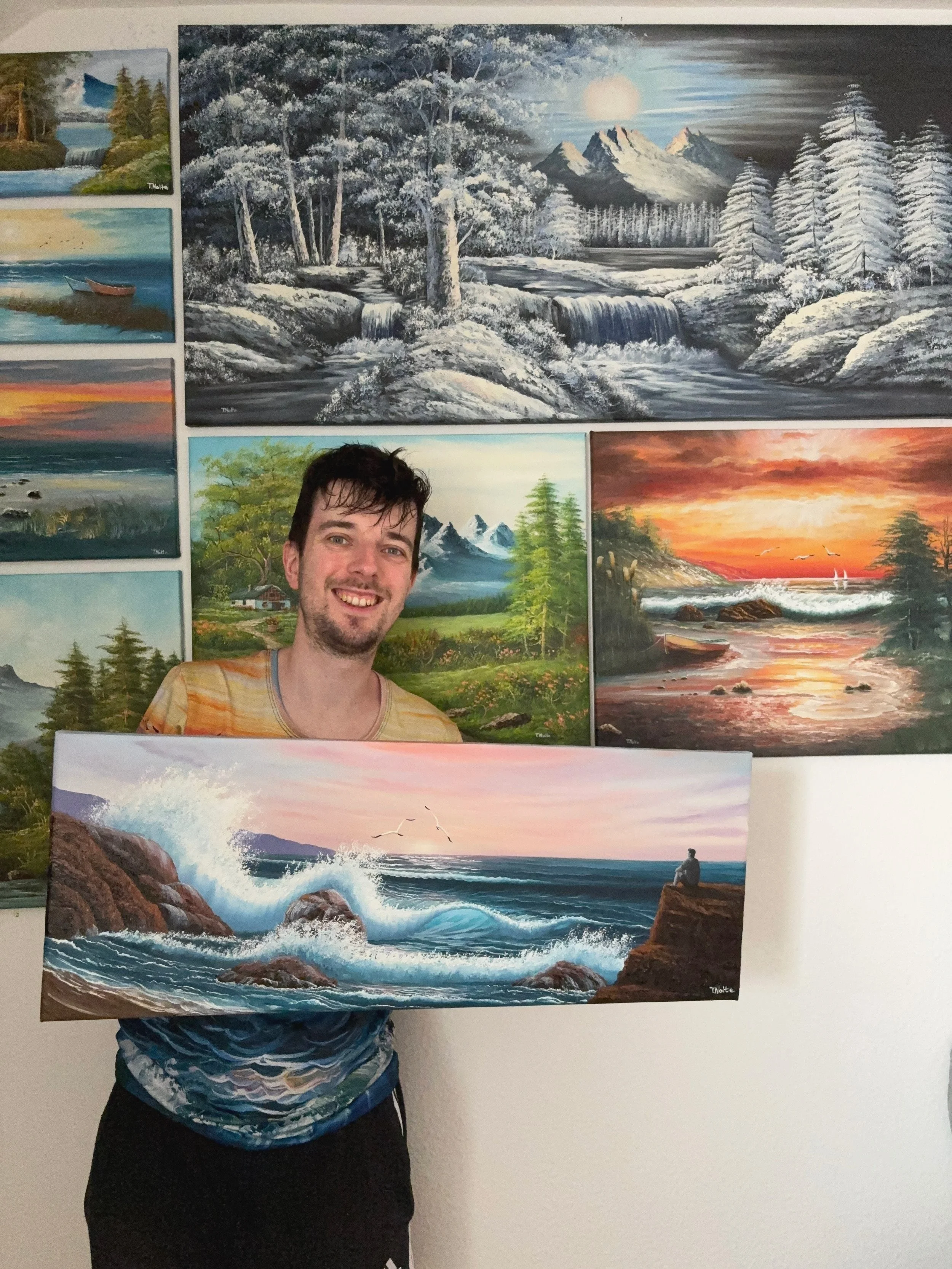A man holding a painting of ocean waves crashing against rocks during a sunset. Behind him, there are several landscape paintings, including mountains, rivers, forests, and sunsets.