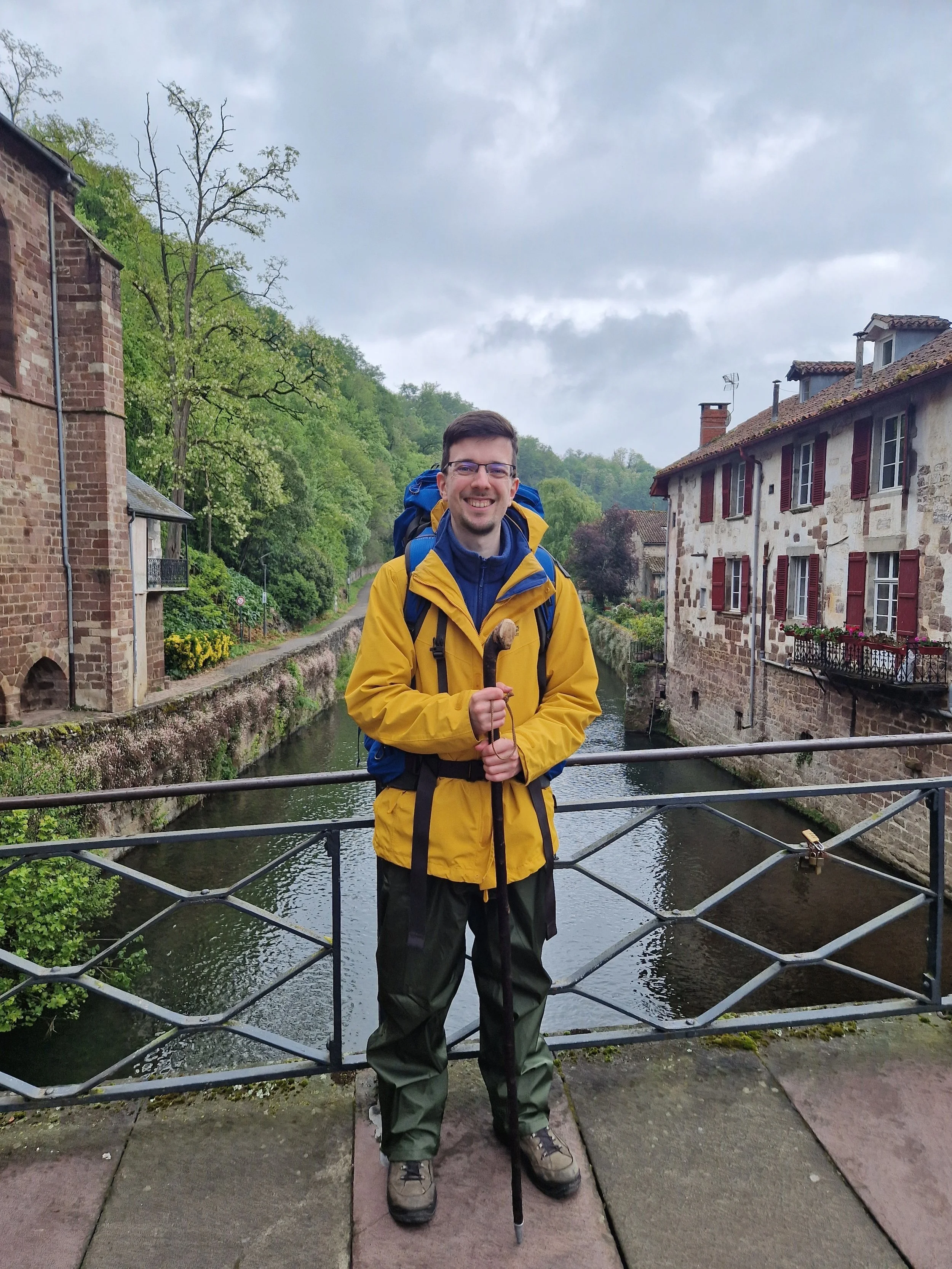 Künstler Tobias Nolte steht auf der historischen Brücke in Saint-Jean-Pied-de-Port, umgeben von Bäumen und fließendem Wasser, als Ausdruck seiner Verbindung zur Natur und Landschaftsmalerei.