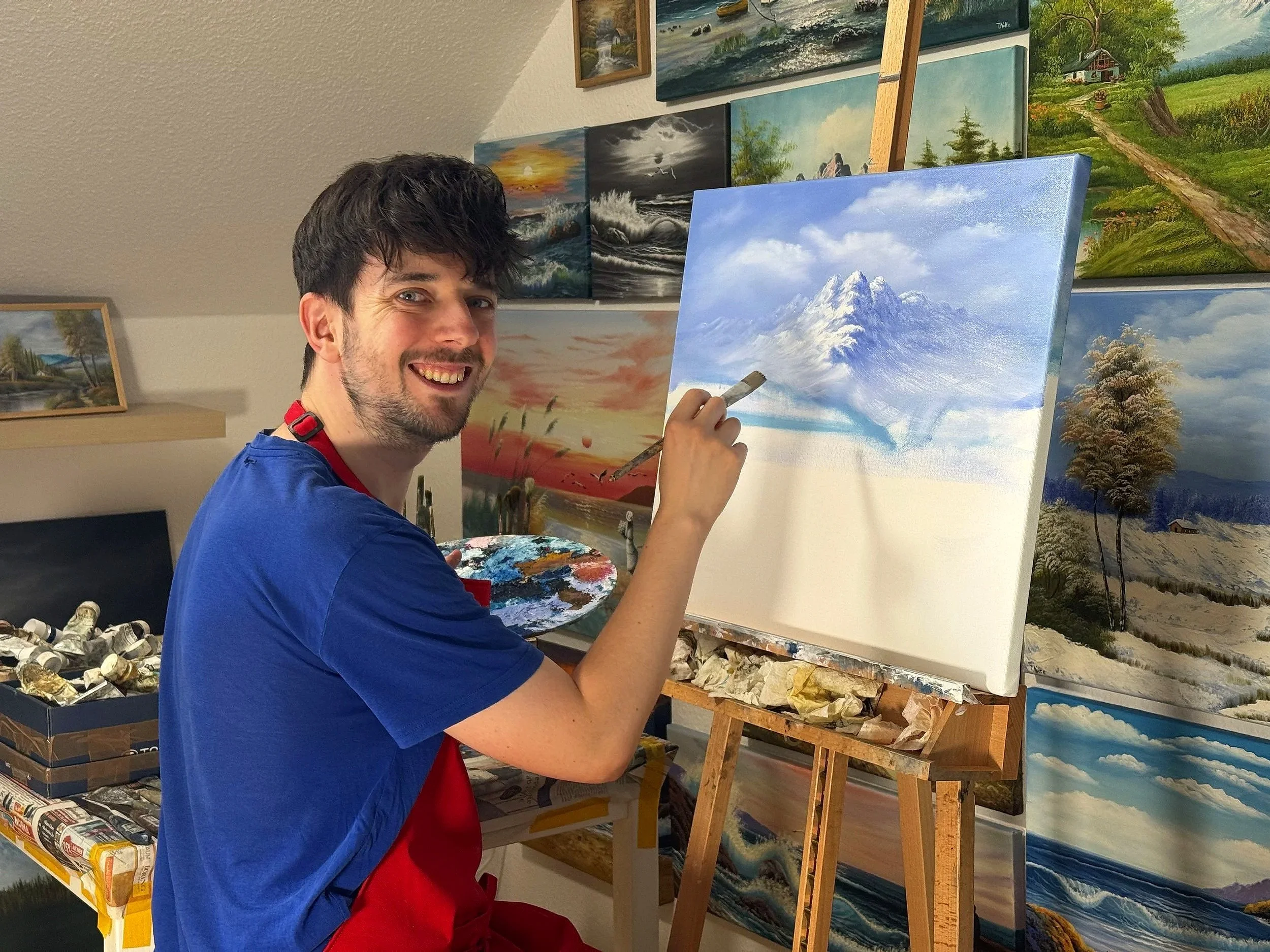 Künstler Tobias Nolte sitzt lächelnd vor einem Landschaftsbild auf Leinwand und malt einen Berg.