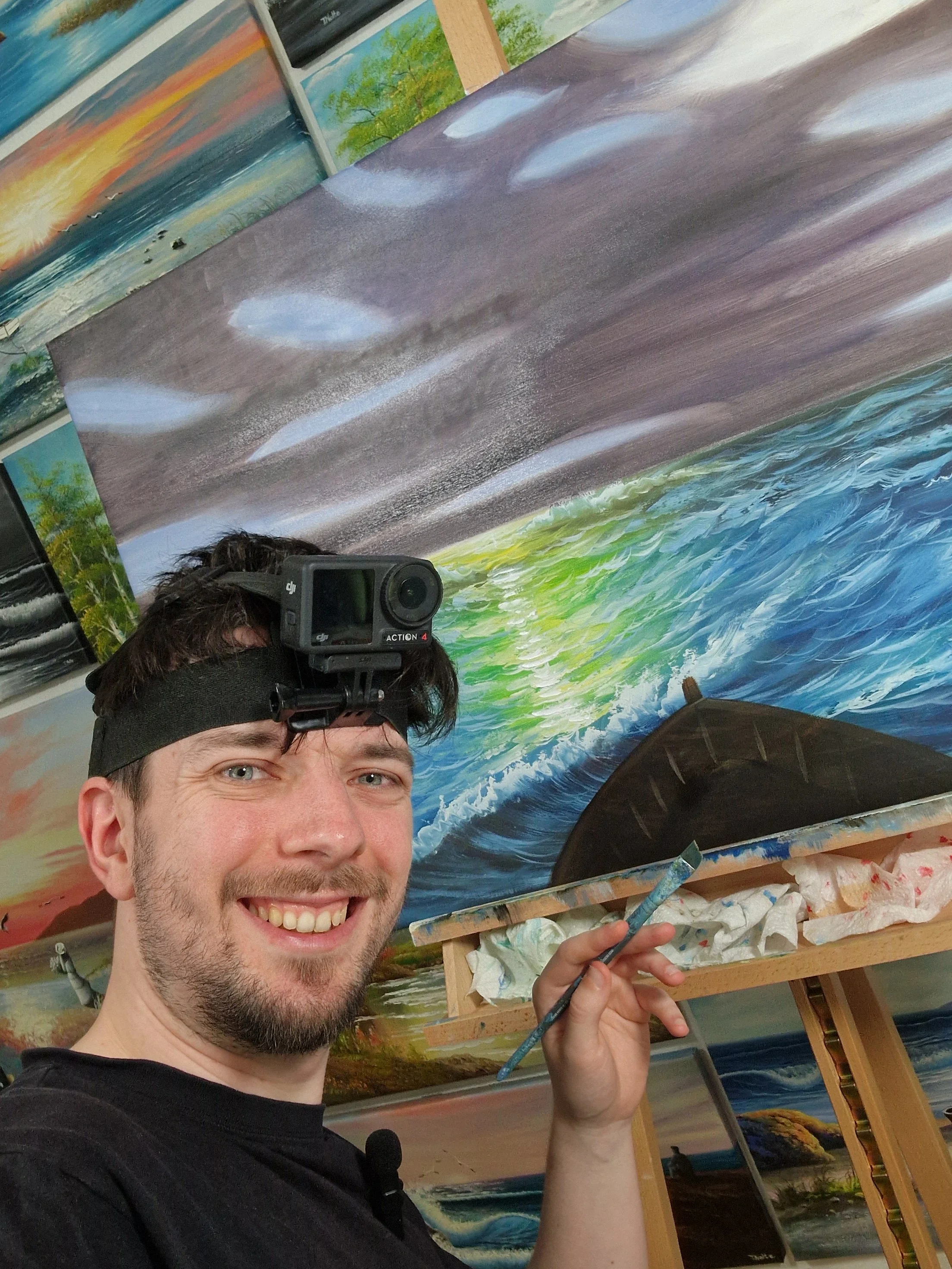 Der Kunststreamer Tobias Nolte mit Helmkamera vor der Leinwand, während der live auf Twitch ein Bild malt.