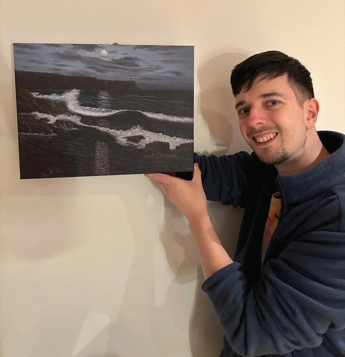Artist holding a small canvas art print of the painting “Night Sea,” featuring moonlit waves and a lighthouse.