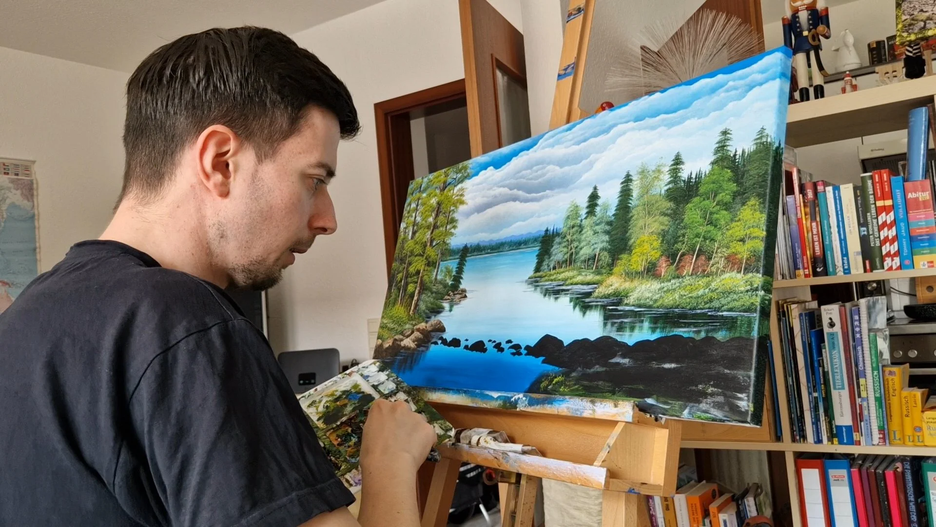 Artist Tobias Nolte sits in front of his landscape painting, adding stone details to a river with a palette knife in hand.