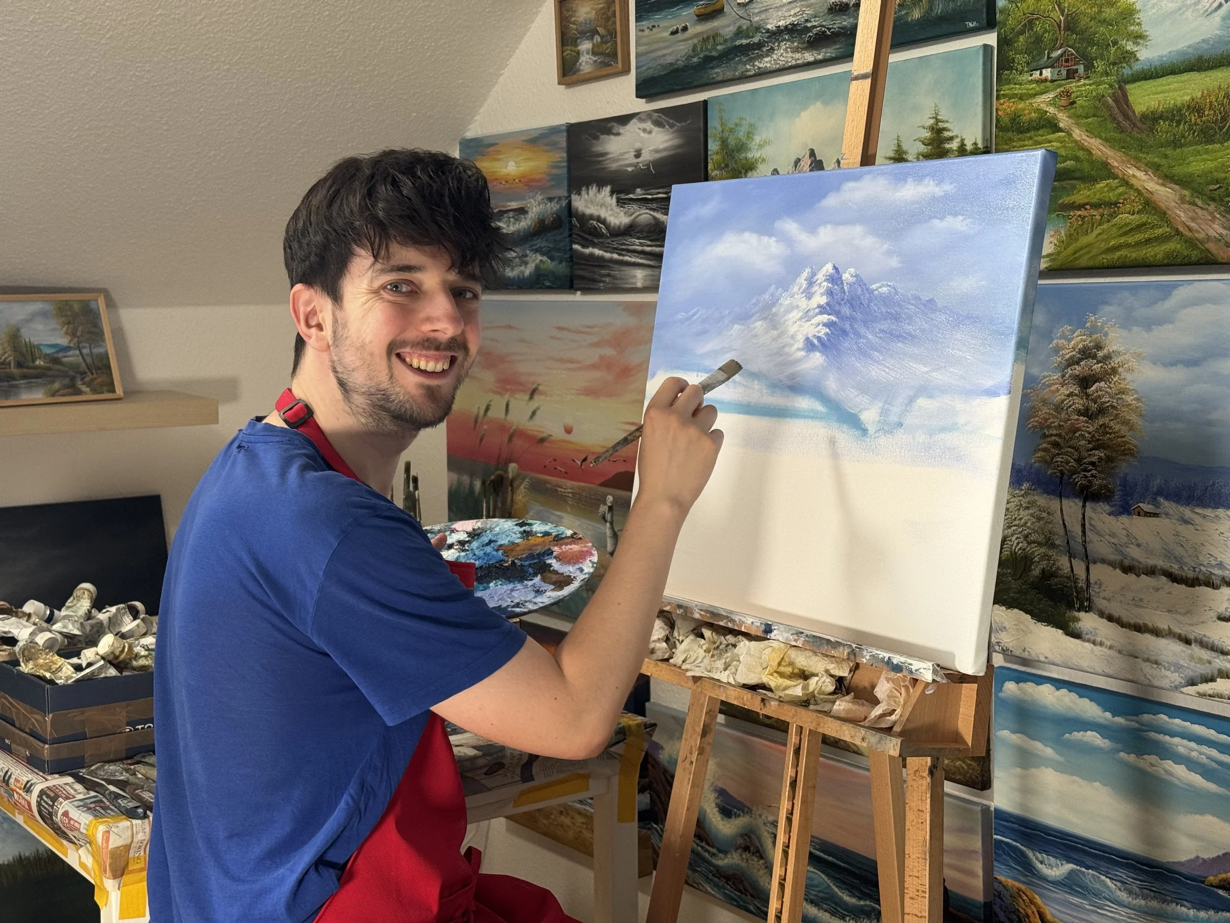 Künstler Tobias Nolte lächelt in die Kamera, während er einen Berg auf Leinwand malt.