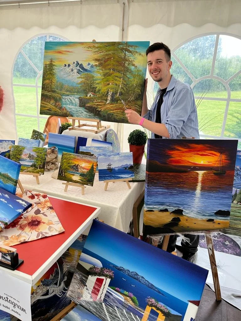 Artist Tobias Nolte stands beside a large painting of a lush green forest with mountains and a waterfall, surrounded by several smaller artworks of his own.