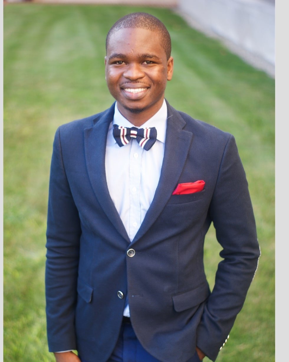 A young man in a dark blue suit with a white shirt, a bow tie, and a red pocket square stands outdoors on grass, smiling at the camera.