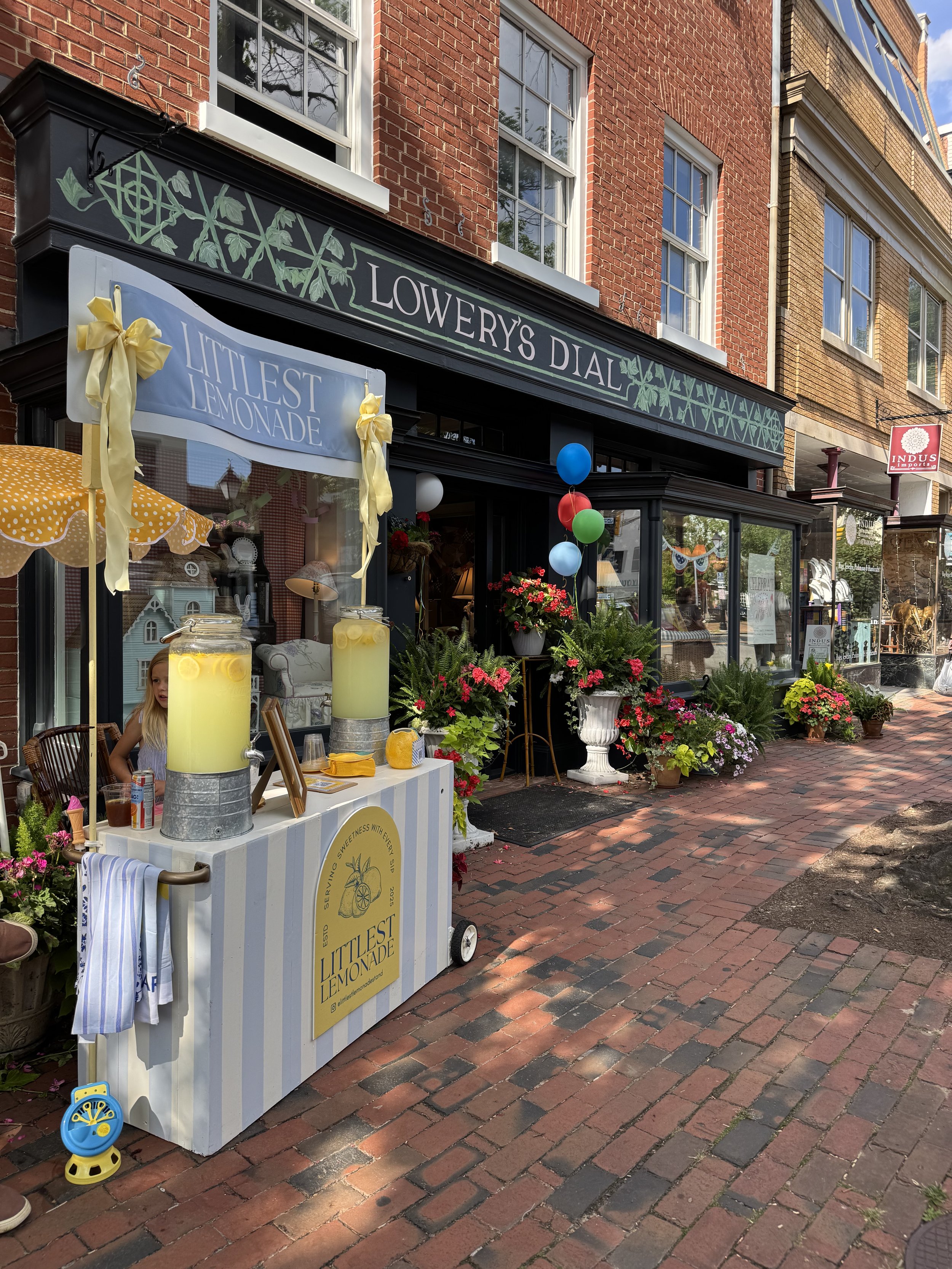 Storefront named Lowery's Dial decorated with balloons and flowers, with a lemonade stand labeled Littleest Lemonade in front, on a brick sidewalk.