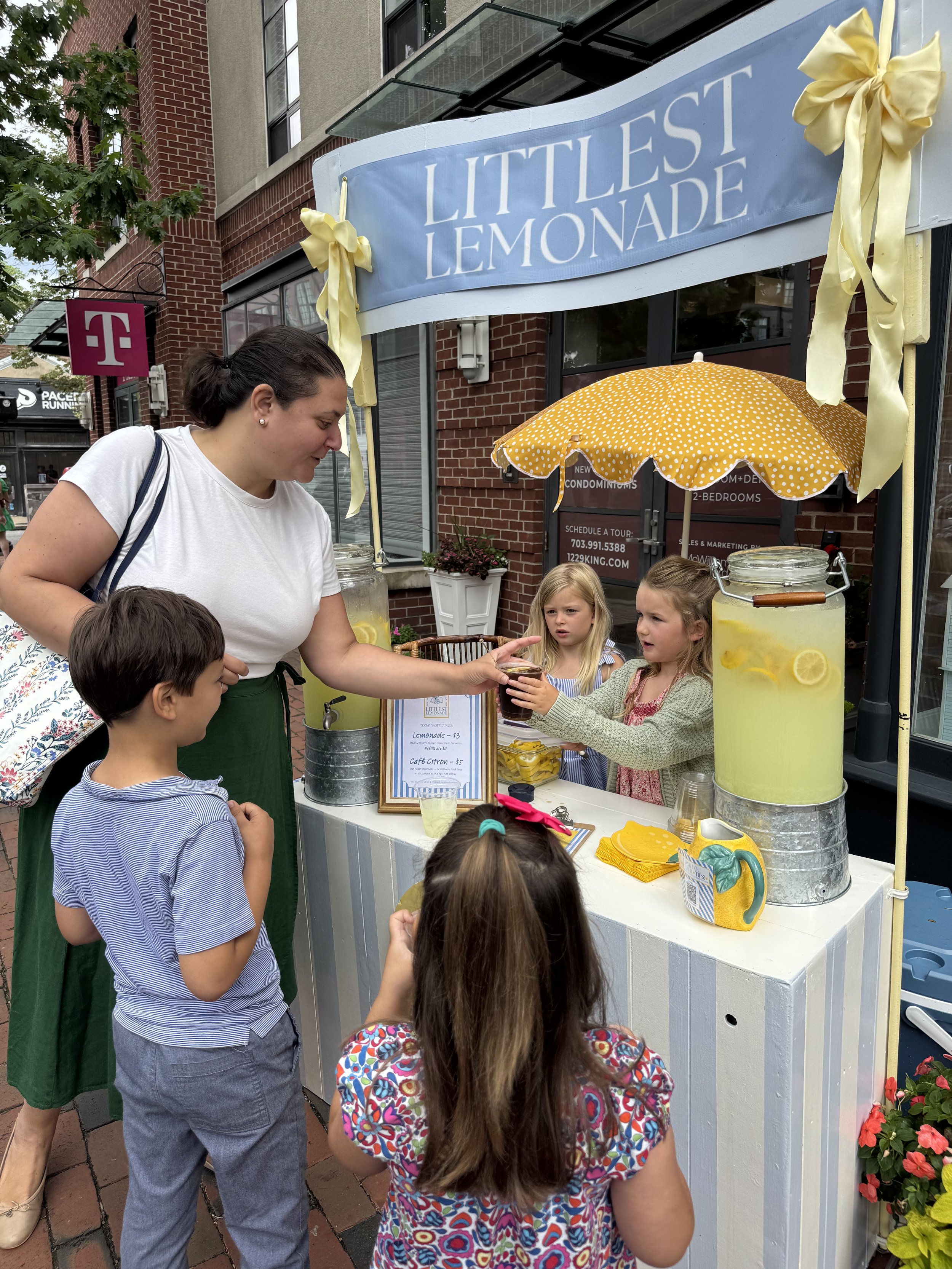 A woman buying lemonade from a stand called 'Littlest Lemonade' with children customers, two girls and a boy, on a sidewalk in front of a brick building, with a yellow polka dot umbrella and large glass jar of lemonade next to the stand.
