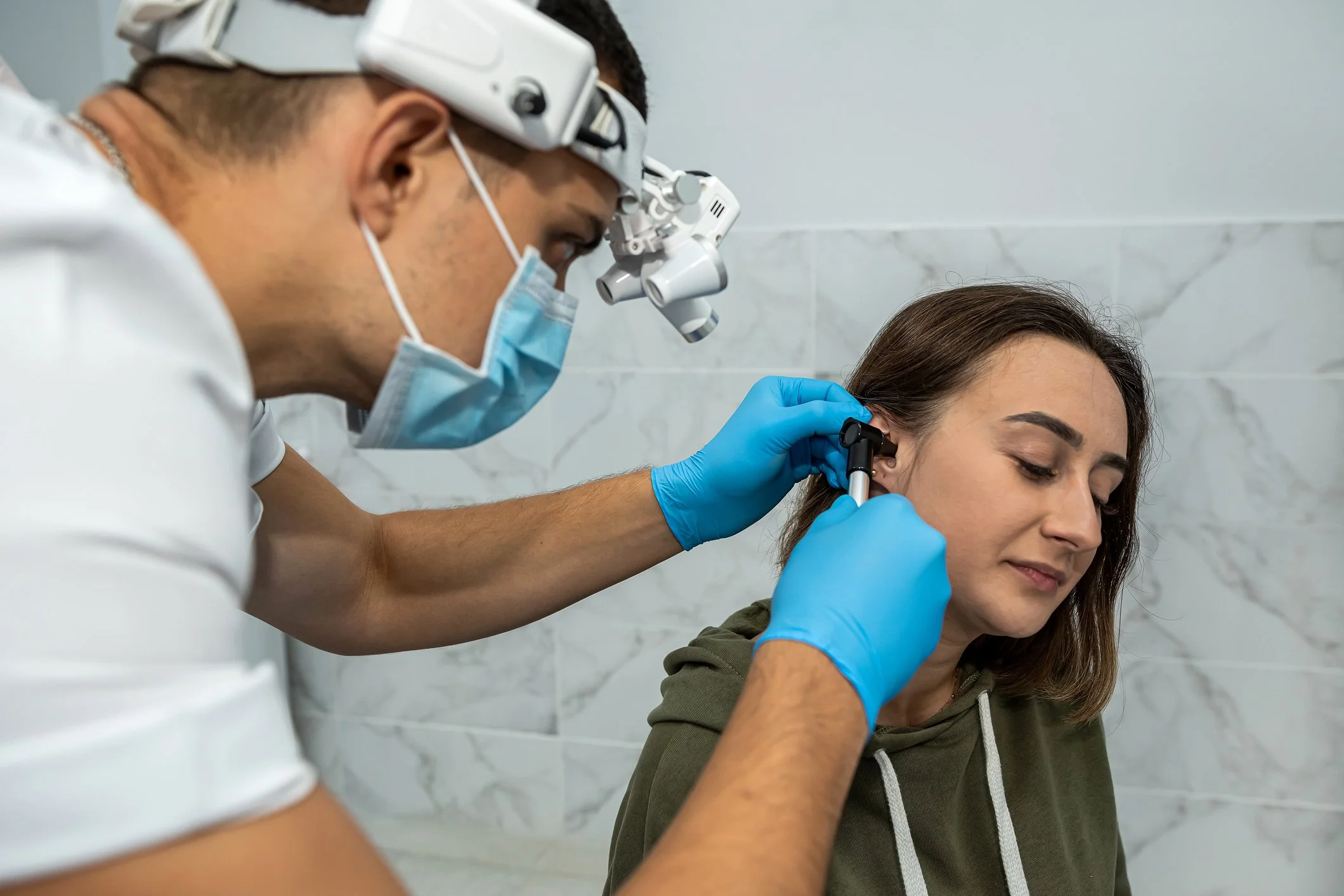 An audiologist examines a woman's ear using a specialized instrument while wearing surgical gloves and a facemask.