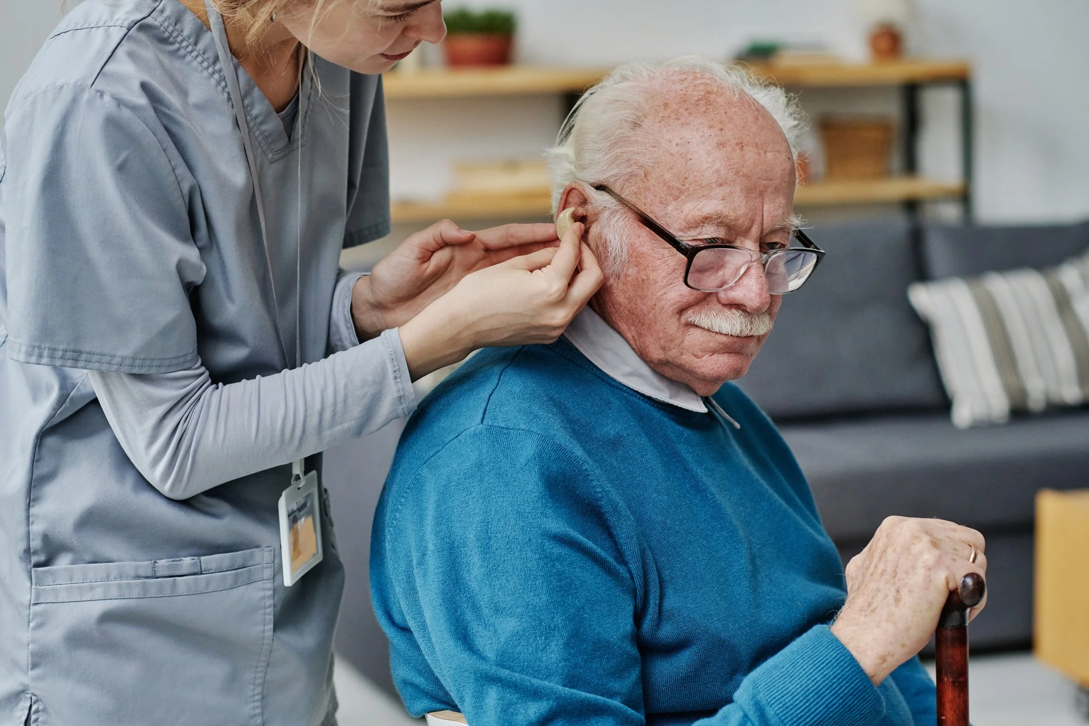 A young female caregiver helping an elderly man with hearing aids in a living room.