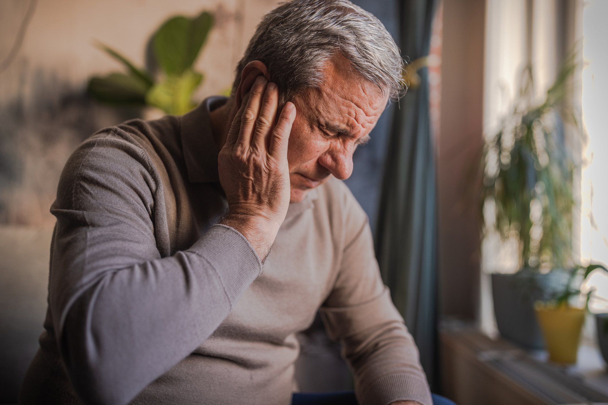 An elderly man is holding his head with his left hand, appearing to experience pain or discomfort, sitting indoors near a window with plants in the background.