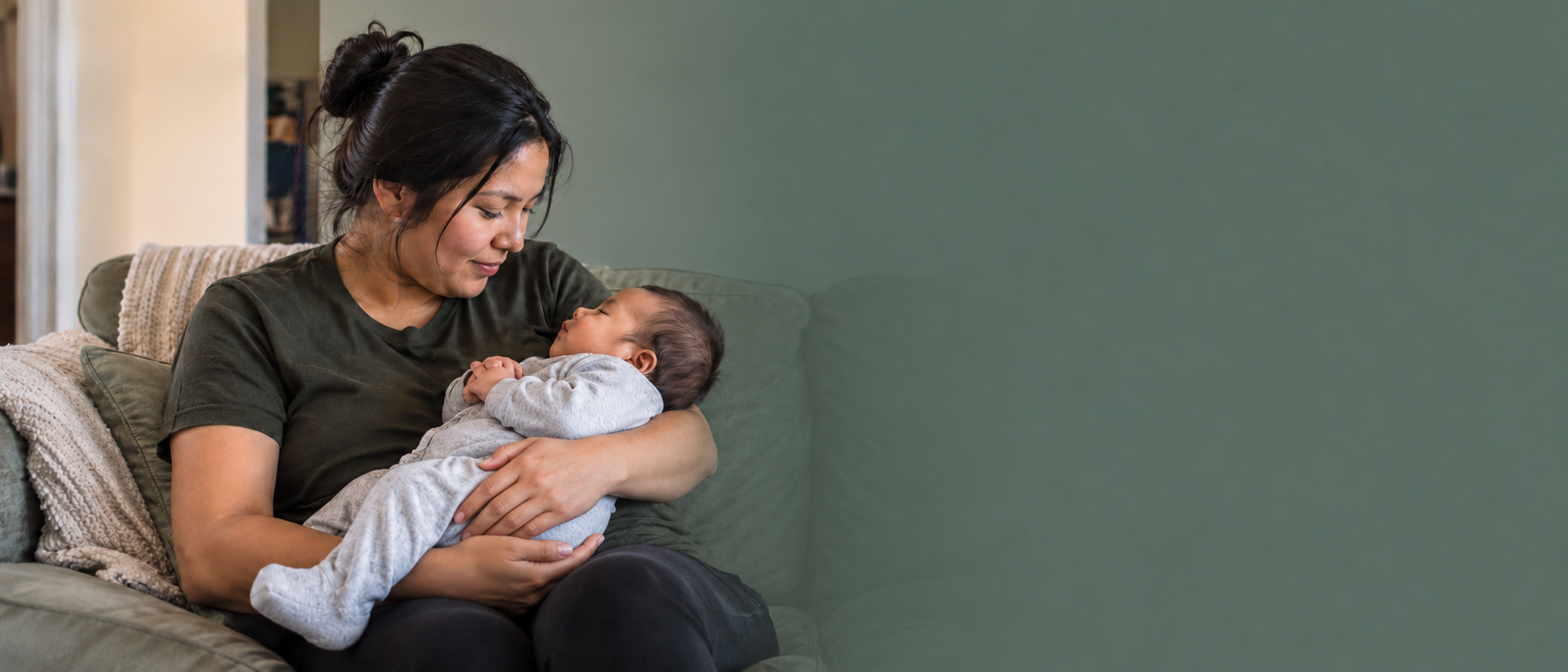 Indigenous mother seated on a sofa, looking down at the infant in her arms.
