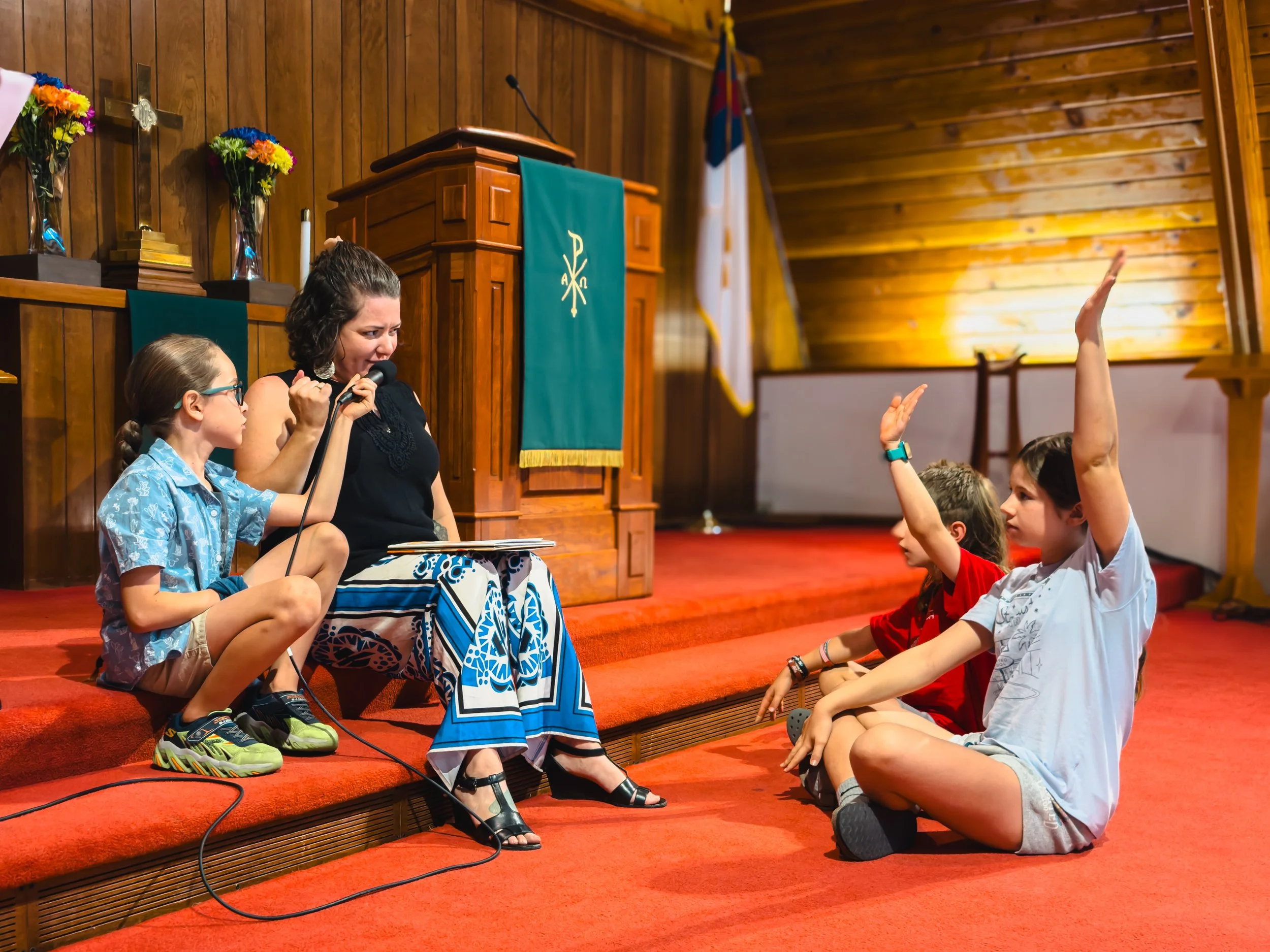 A woman and child sit on a step in a church, with two kids across from them with their hands raised.