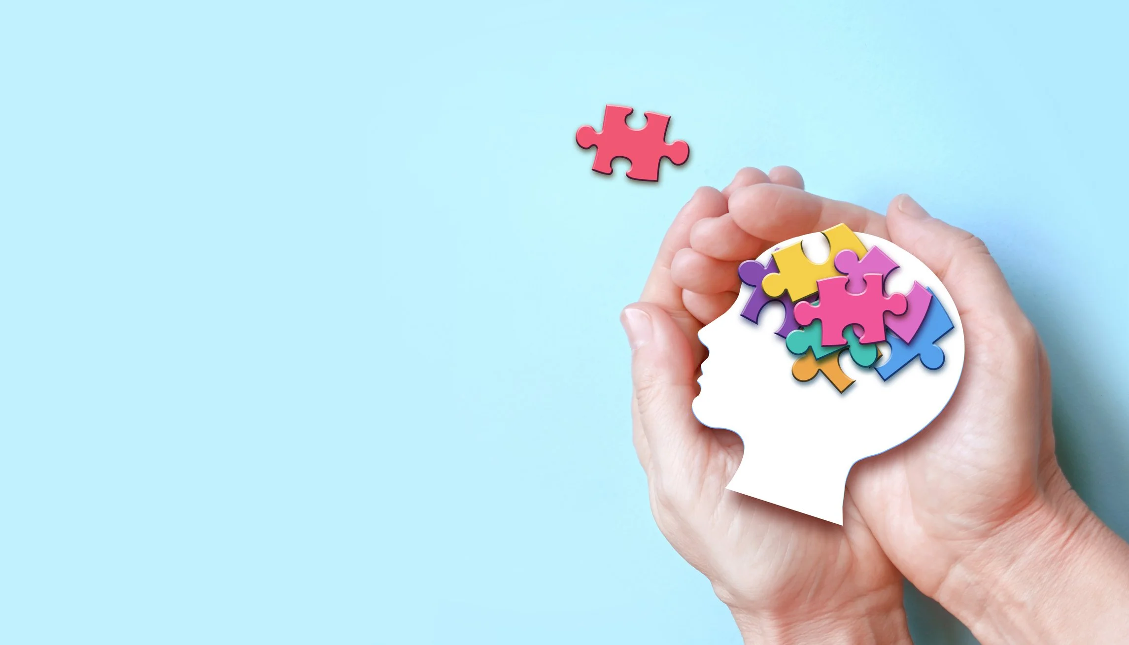 A person's hands holding a white puzzle piece in the shape of a human head, filled with colorful puzzle pieces representing a brain, with one puzzle piece floating above against a light blue background.