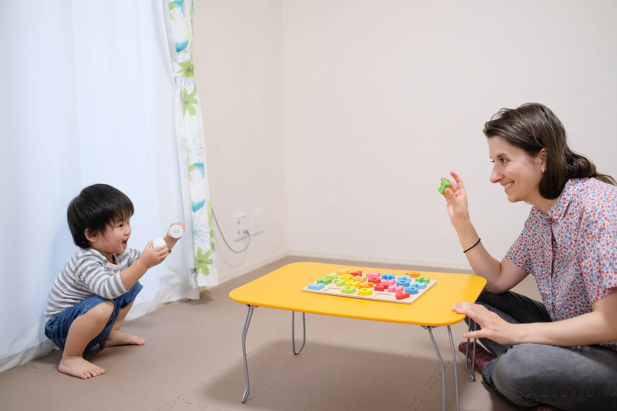 A woman and a young boy playing a game with colorful wooden alphabet puzzles at a small yellow table in a room with beige carpet and white walls.