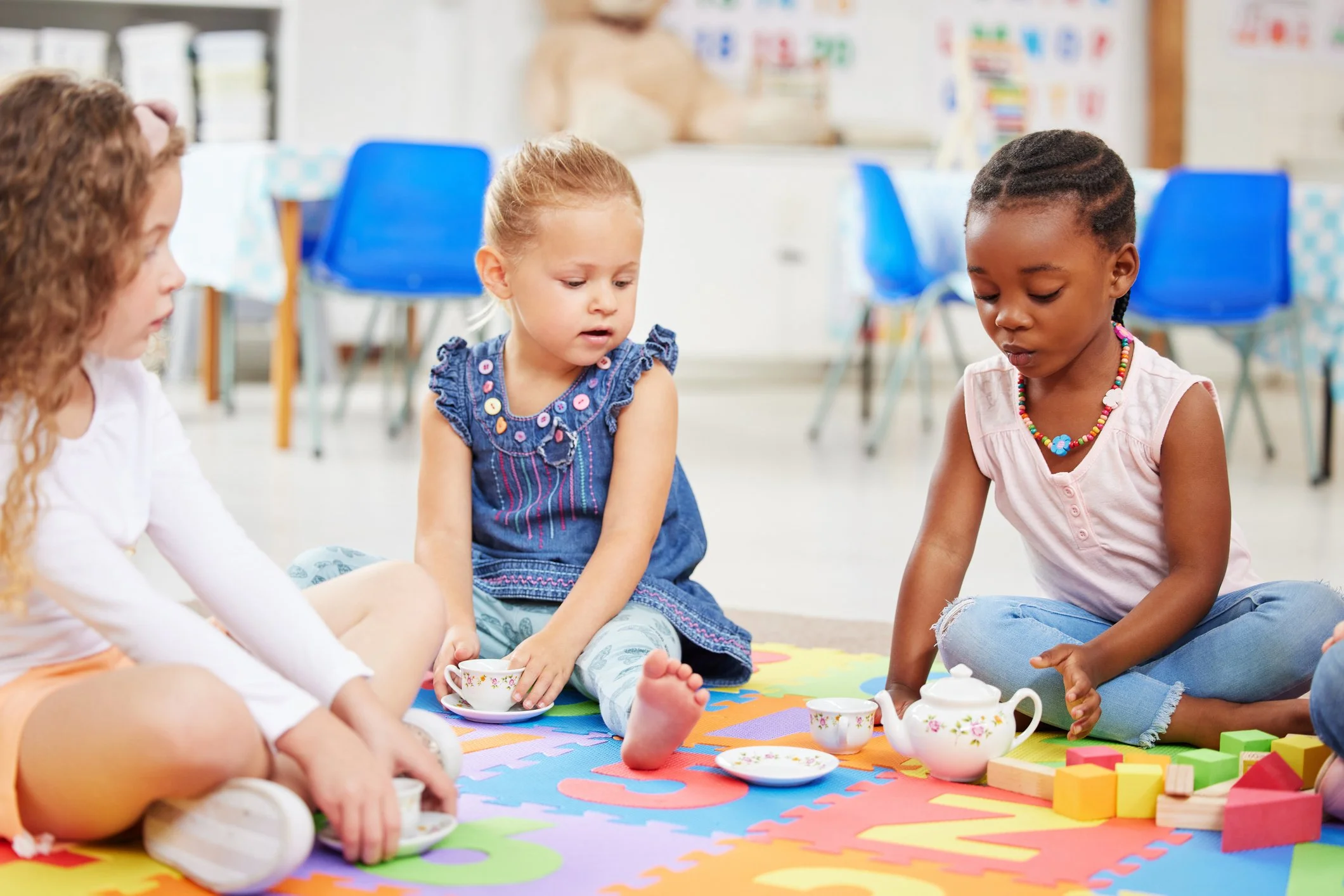 Three young girls sitting on a colorful foam mat in a classroom, playing with toy tea set and wooden blocks.