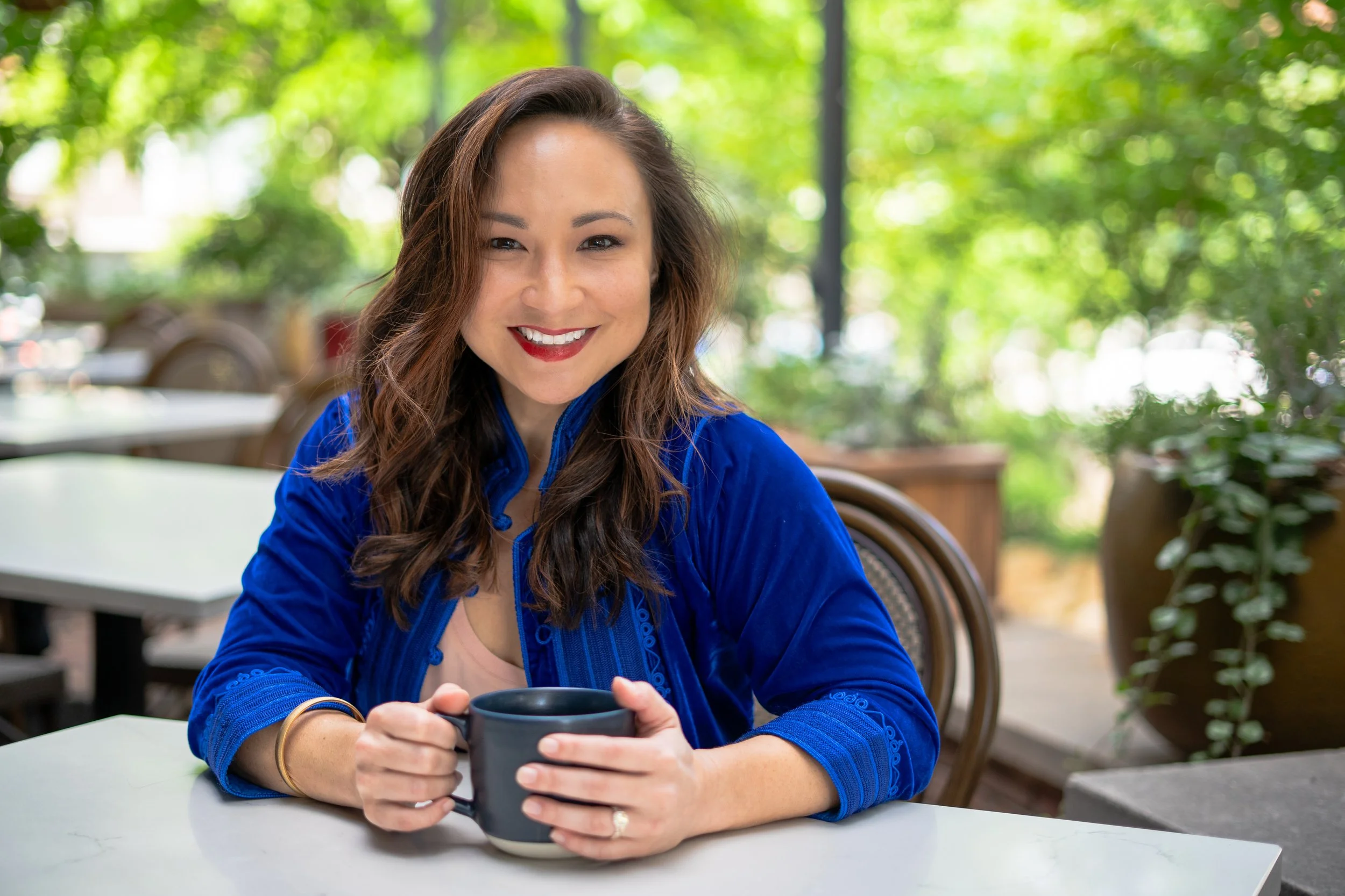 A woman with long wavy hair smiling while sitting at a table holding a black coffee mug, in an outdoor setting with green foliage in the background.