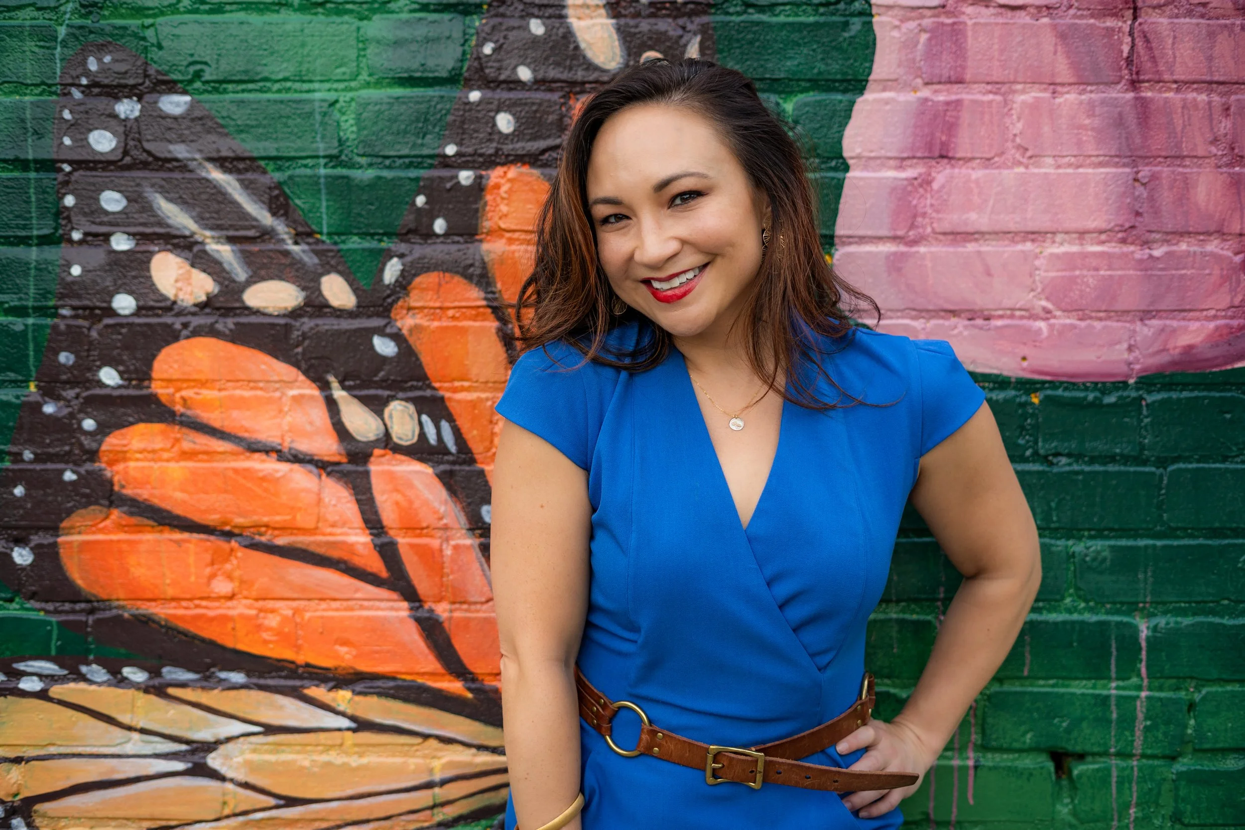 A woman in a blue dress smiling in front of a colorful wall mural of a butterfly and flowers.