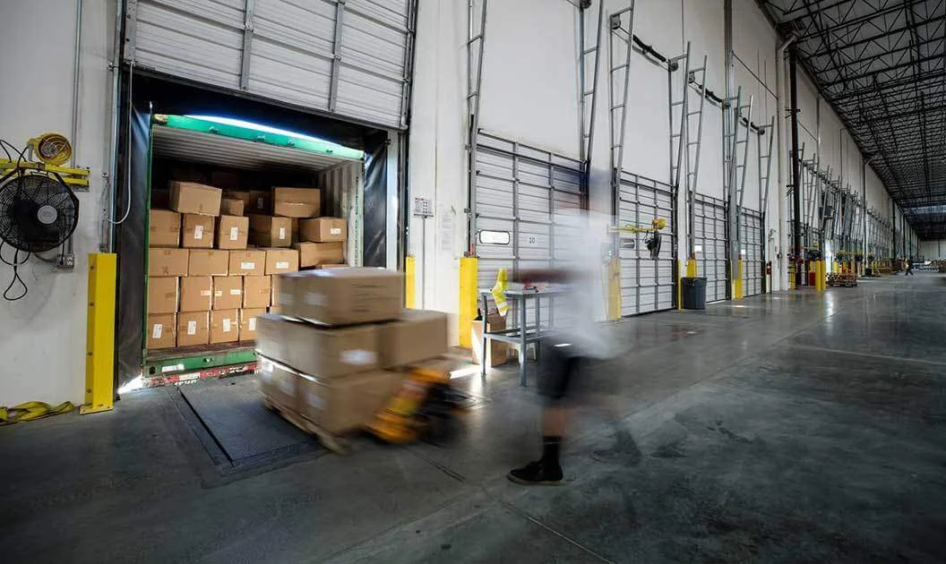 A worker in motion moving a dolly loaded with cardboard boxes into a warehouse storage area. The warehouse has a high ceiling, multiple large garage doors, and industrial equipment along the walls.
