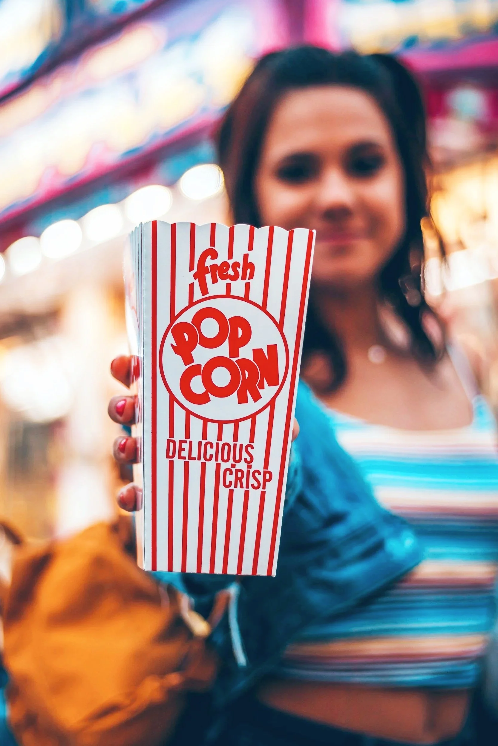 A woman holding a large striped red and white popcorn container at a fair or carnival, with colorful lights in the background.