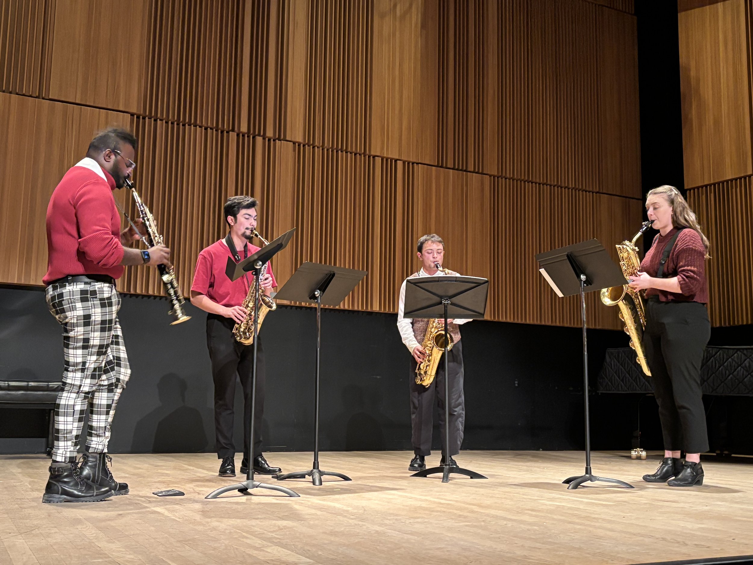Four musicians playing saxophones on a stage with wooden paneling.