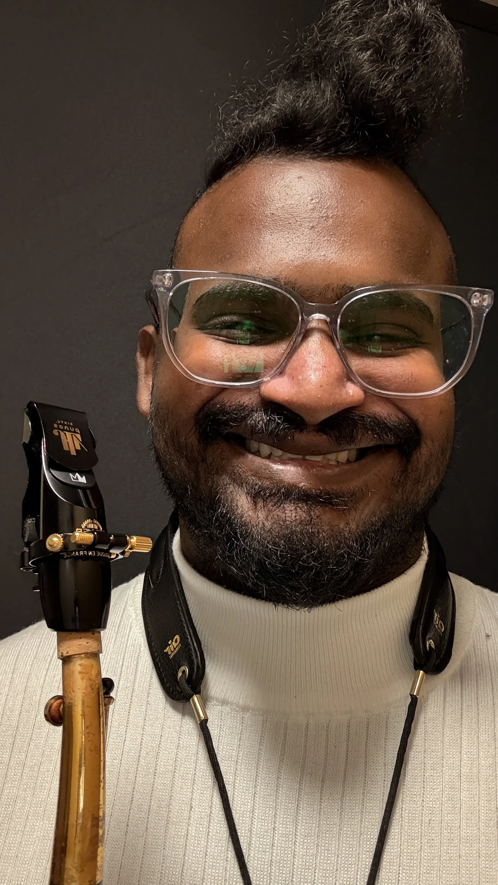Close-up of a smiling man with glasses, dressed in a white turtleneck, carrying a camera strap around his neck, holding a wooden cane, with a dark background.