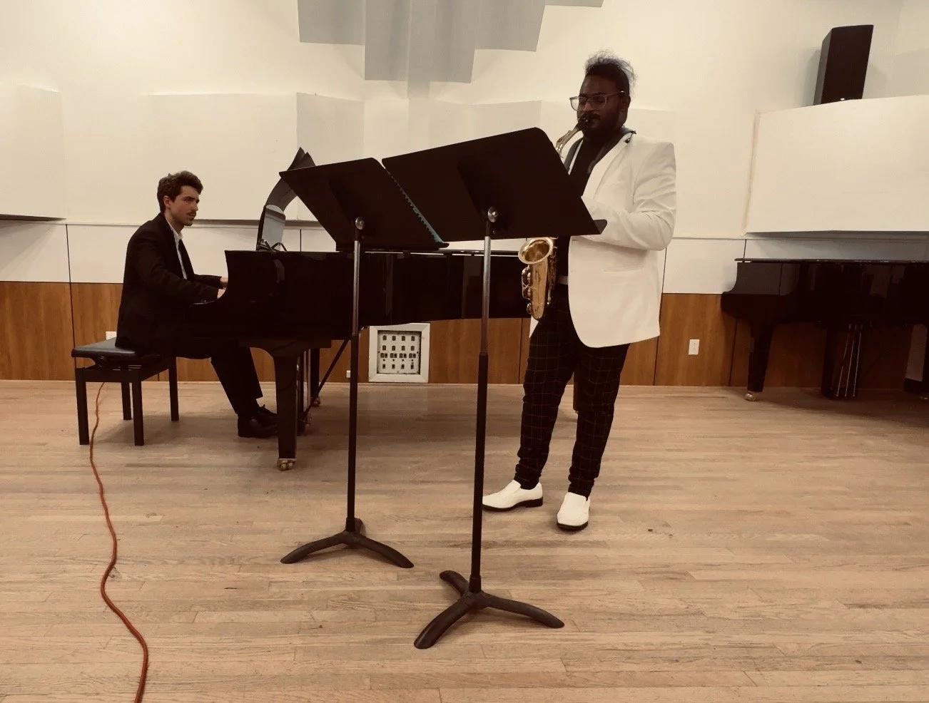 A musician in a white blazer playing the saxophone and a pianist in a black suit playing the piano in a music rehearsal room.
