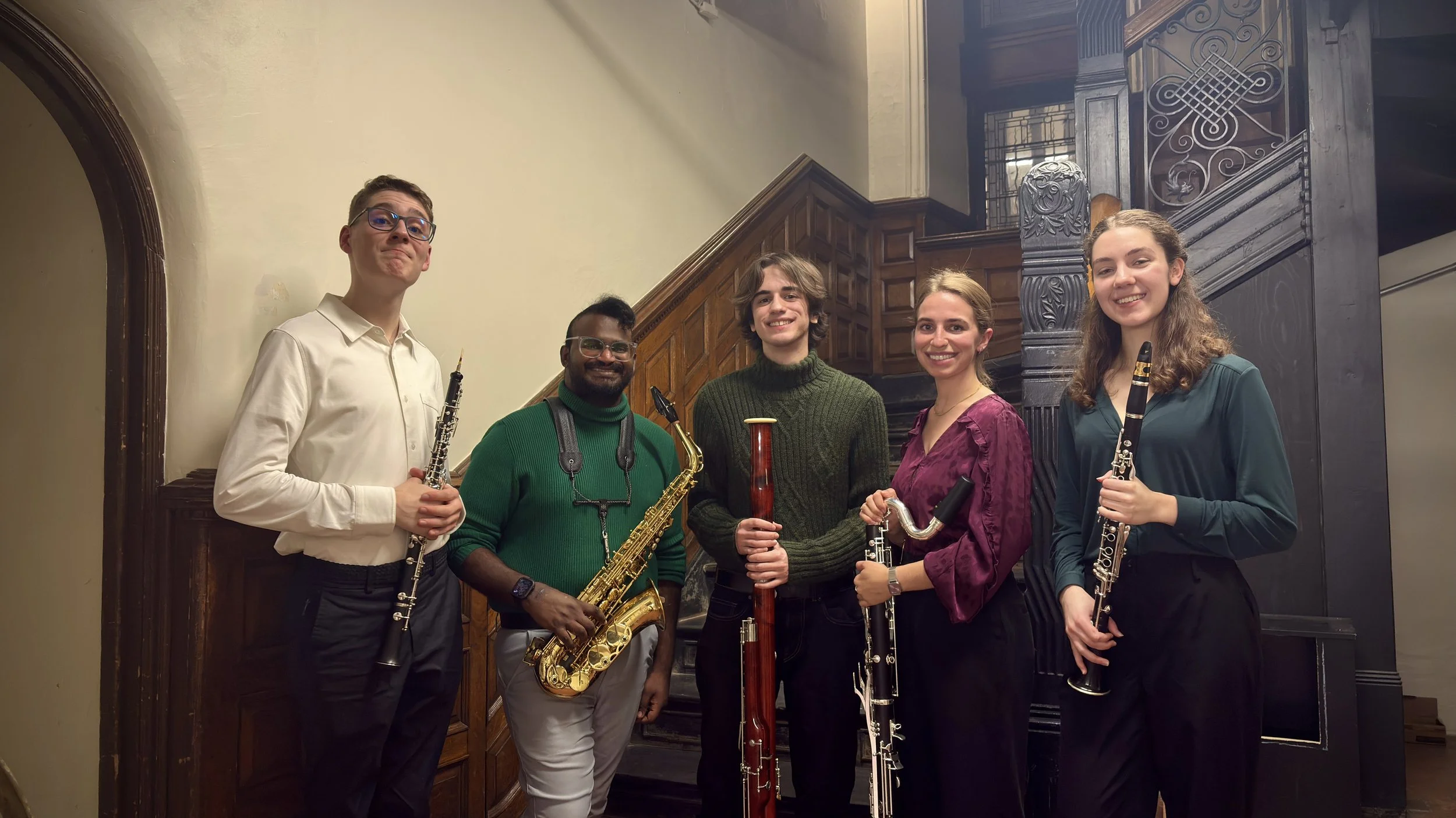 Group of five young musicians holding woodwind instruments in a vintage indoor setting with dark wood staircase.