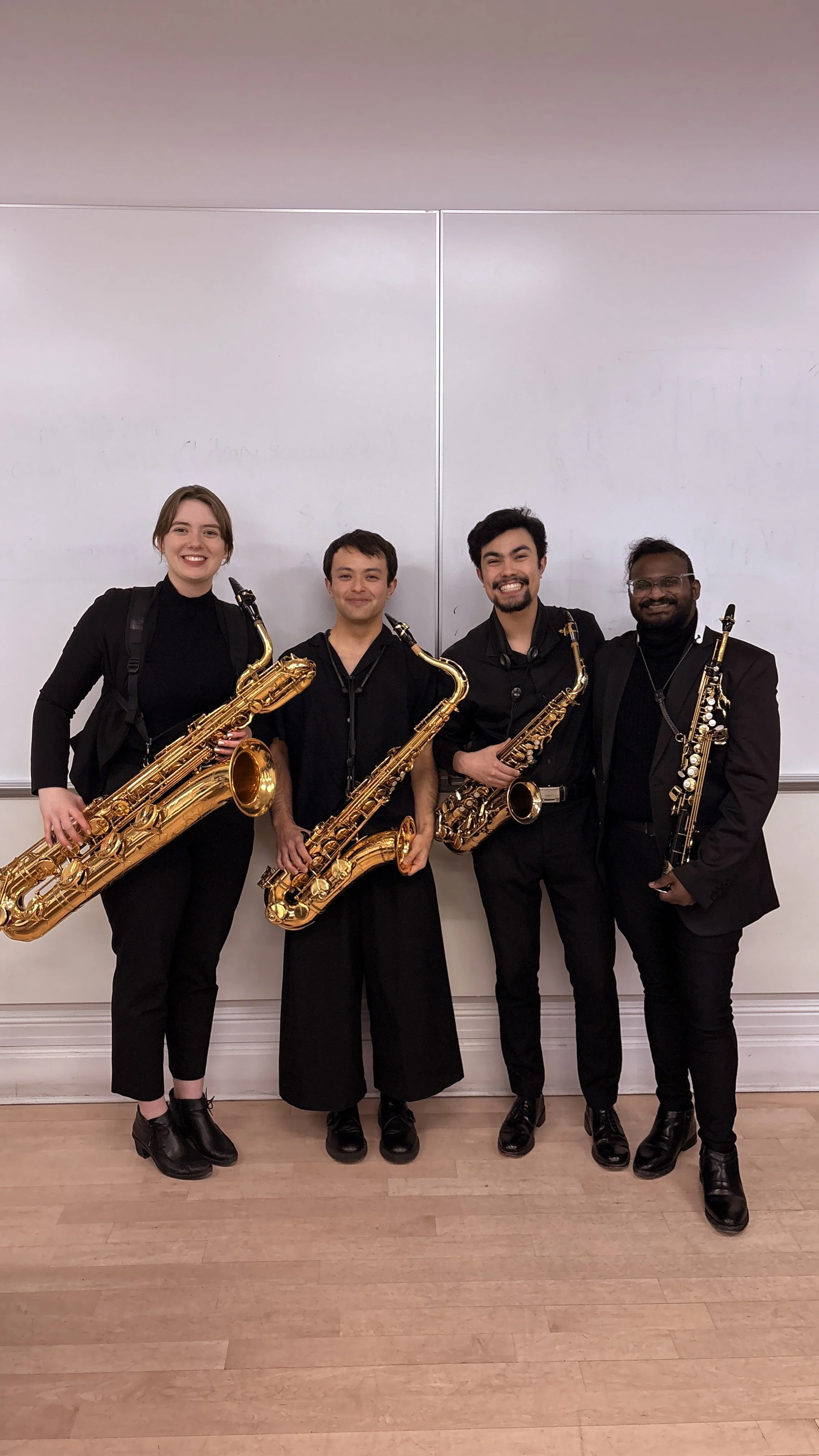 Four young musicians in black clothing, each holding a woodwind instrument, standing in front of a whiteboard in a classroom or rehearsal room.