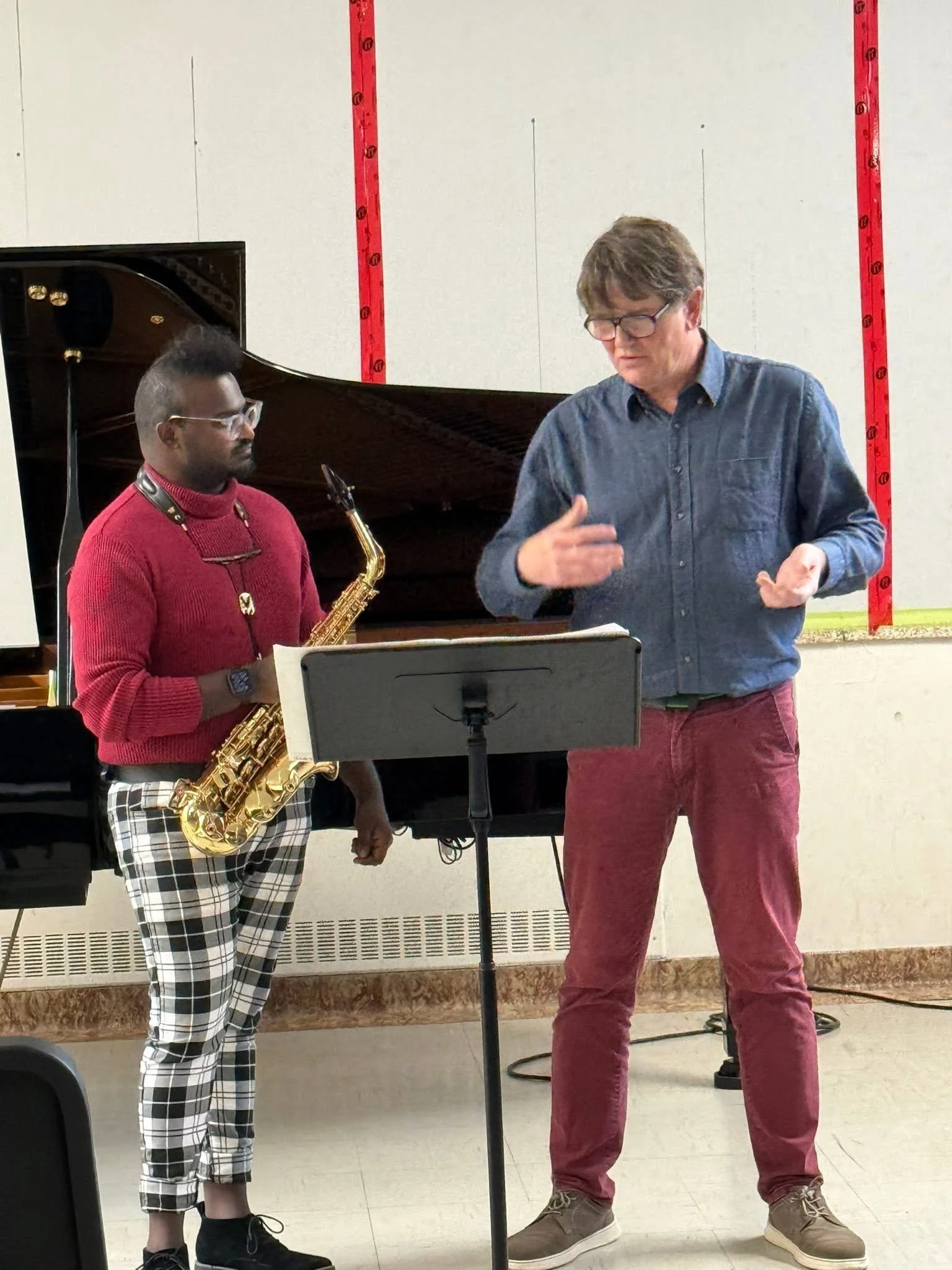 A music instructor and a student engaged in a music lesson in a classroom. The student holds a saxophone, and both are standing near a music stand. There is a piano in the background.