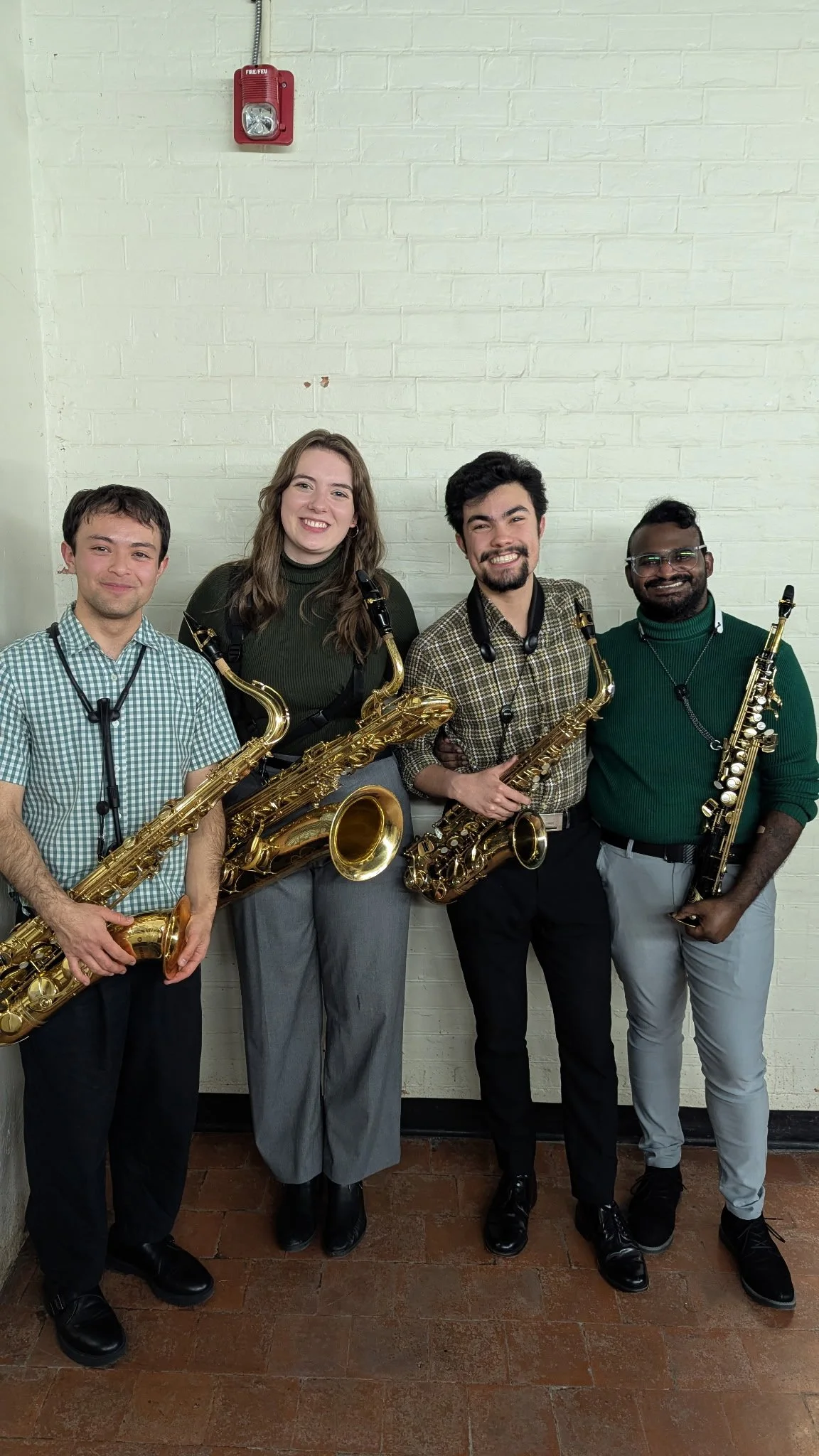 Four musicians holding saxophones, standing in front of a white brick wall, smiling.