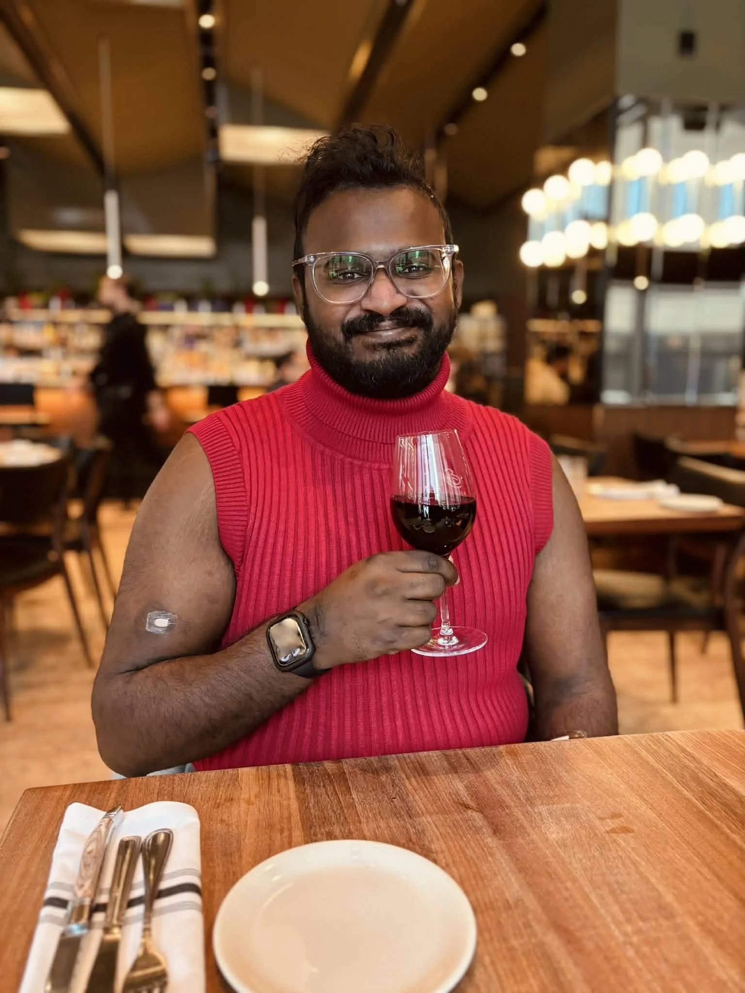 A man with glasses and a beard wearing a red sleeveless turtleneck sweater, holding a glass of red wine, seated at a restaurant table with silverware and a small plate in front of him.